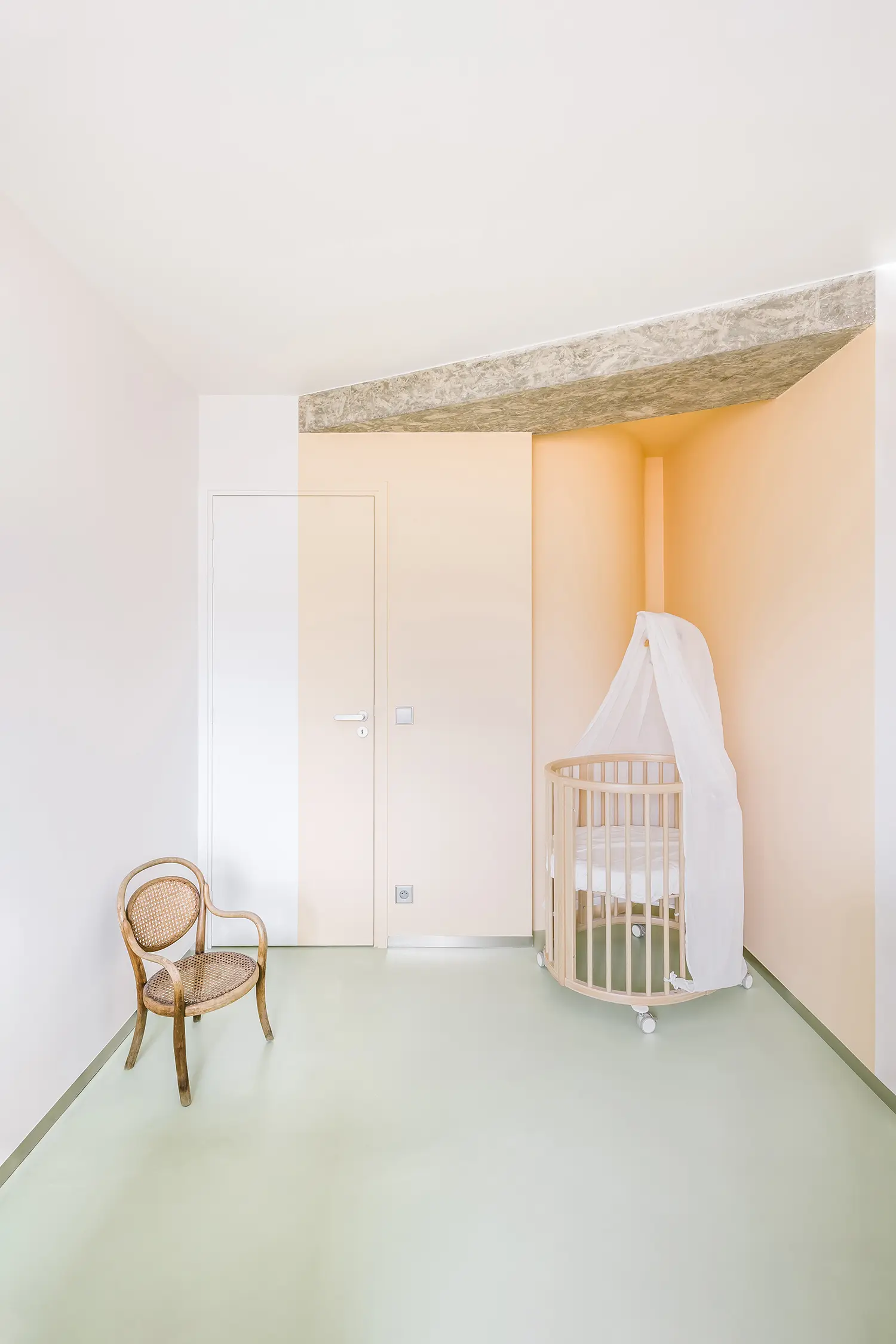 A child's nursery with a peach-colored accent wall, a wooden crib, and an exposed raw concrete ceiling beam.