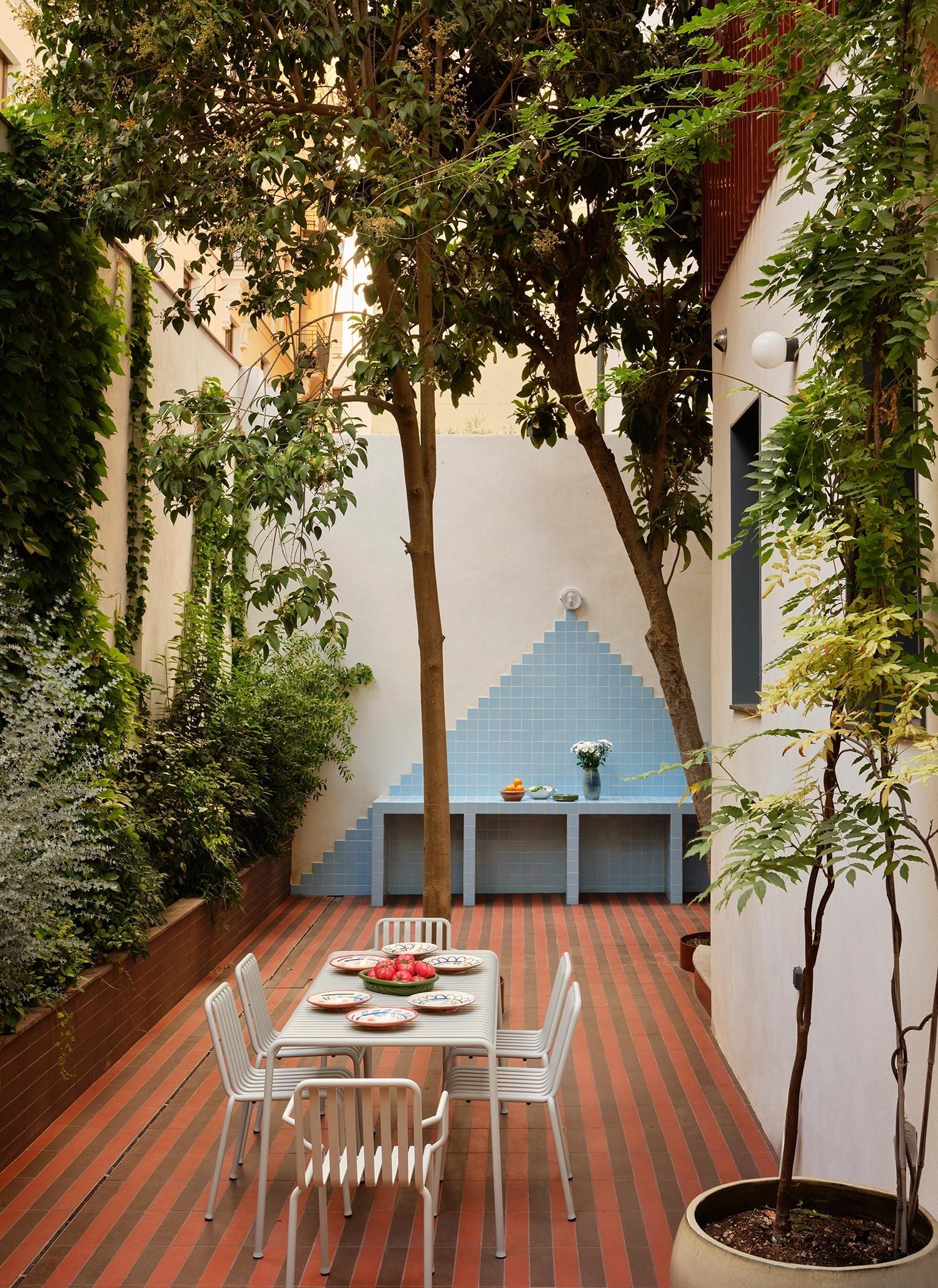 A compact urban patio area with striped red tile flooring, dark wooden outdoor lounge furniture, concrete steps, and a lush vertical wall covered in climbing green ivy.