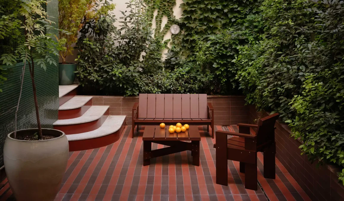 High-angle view of a narrow urban courtyard with a light blue tiled outdoor kitchen counter, striped red and brown flooring, and white tubular metal furniture under green trees.