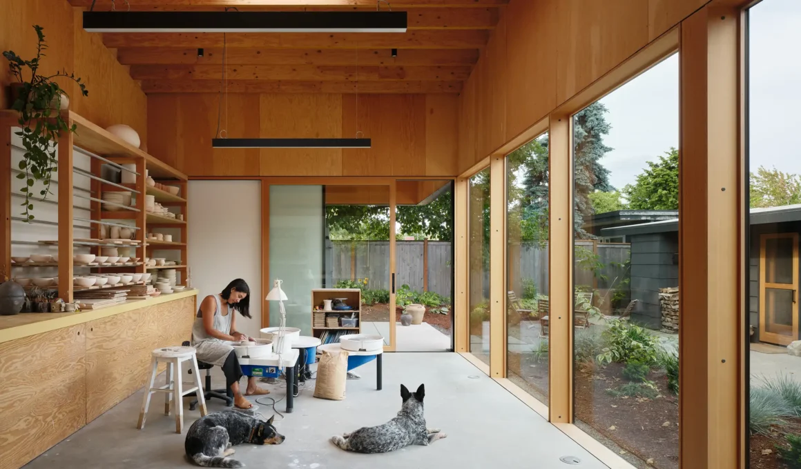 Ceramicist working at a pottery wheel in a sun-drenched timber studio with two dogs resting on the concrete floor.