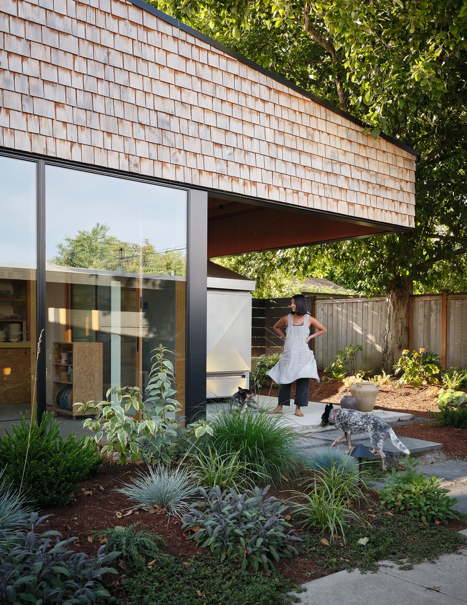Exterior view of a cedar-shingled DADU with a large cantilevered roof and glass walls overlooking a landscaped garden.