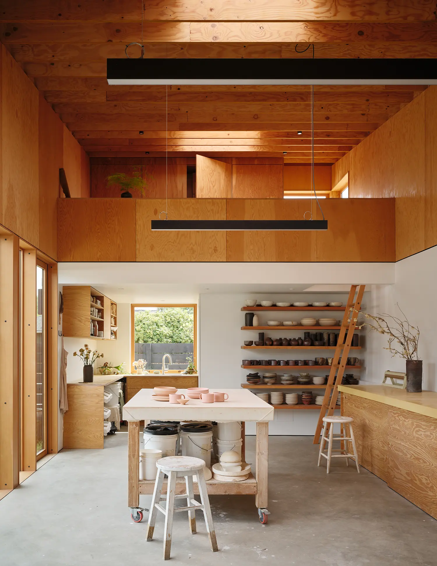 Double-height interior of a ceramics workshop featuring a loft, exposed timber rafters, and a central work table.