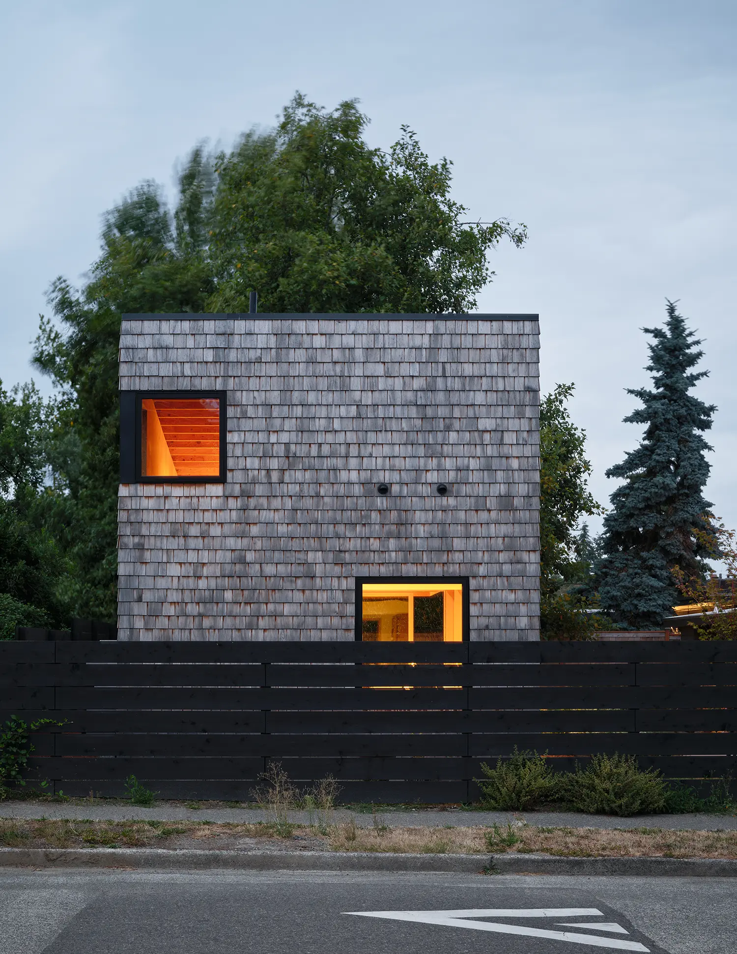 Evening view of a cedar-shingled studio facade with warm light glowing through square windows behind a black fence.