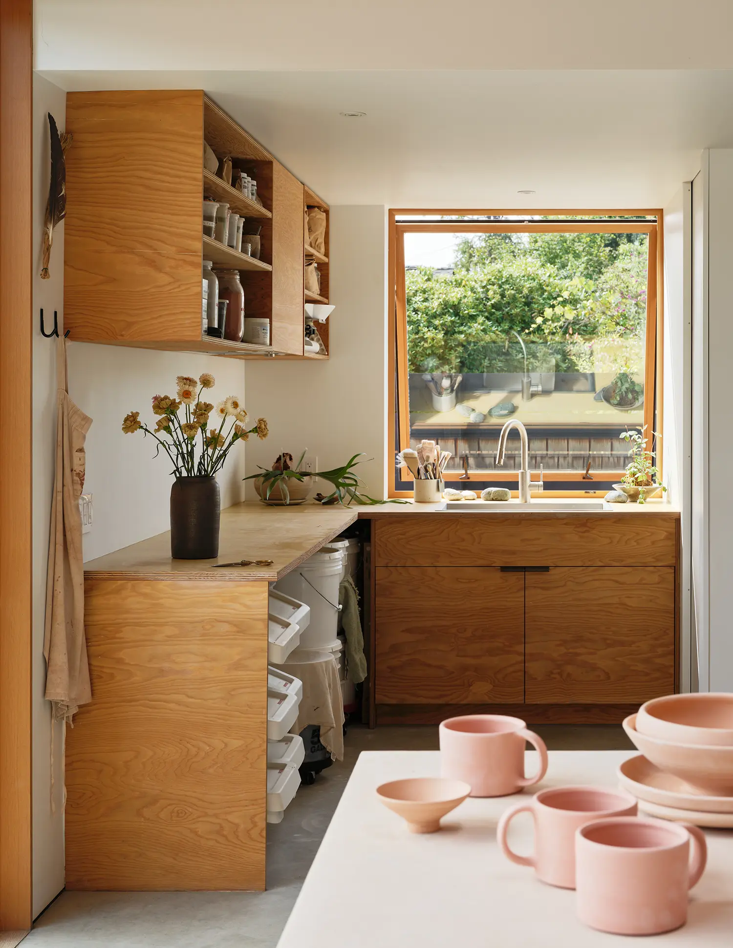 Close-up of a studio kitchen area with plywood cabinets, a large window viewing the garden, and pink ceramic mugs in the foreground.