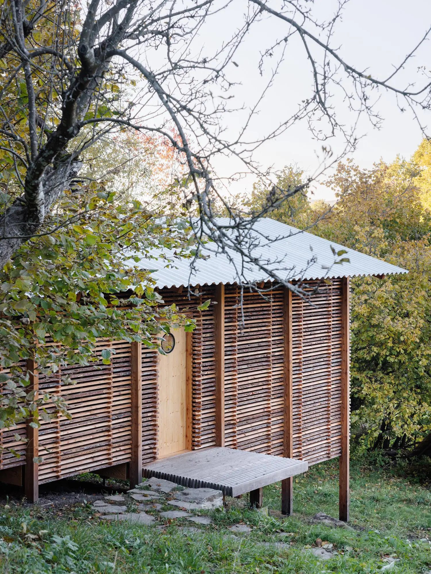 Wooden walkway leading to an entrance door with a small porthole window on a slatted timber building facade.