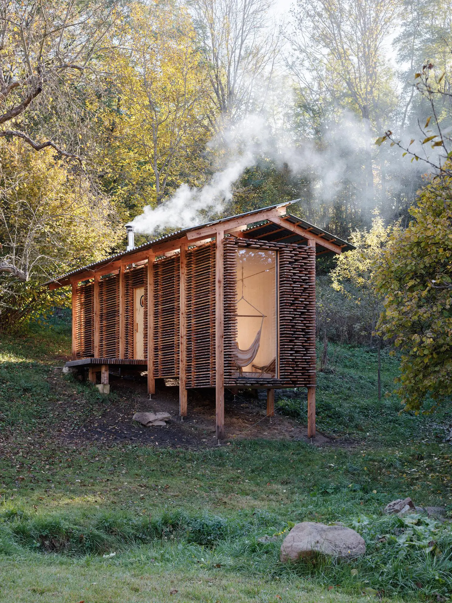 Slender wood sauna building on a slope with smoke rising from a metal chimney amid autumn foliage.