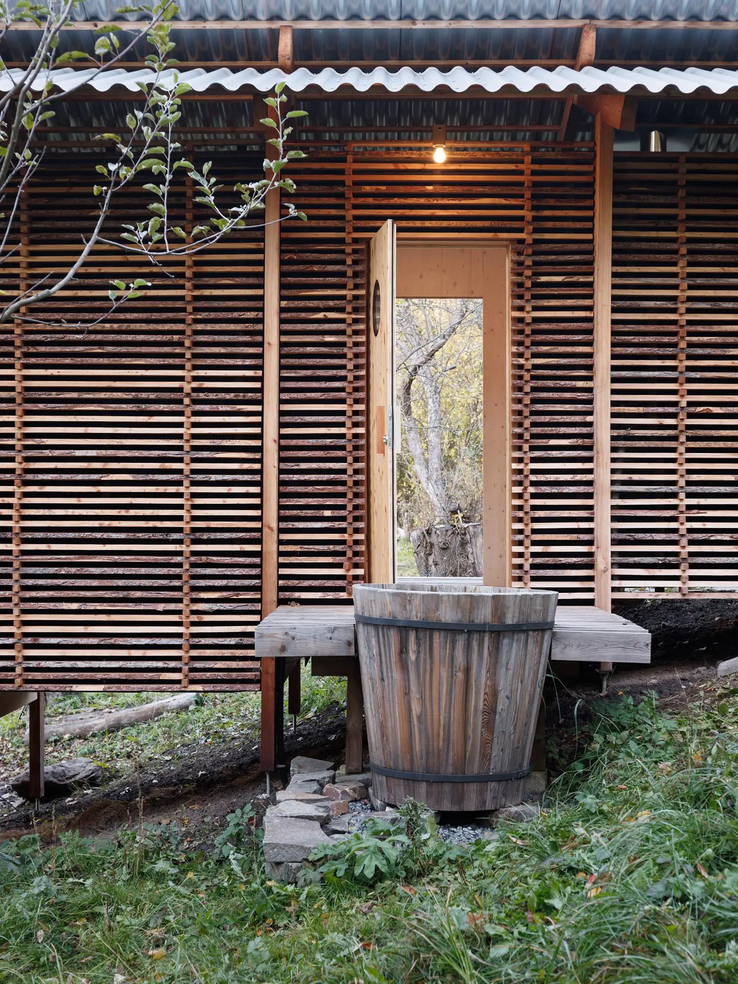 Outdoor wooden terrace with an open door and a round timber cooling tub placed on stone footings on a hillside.