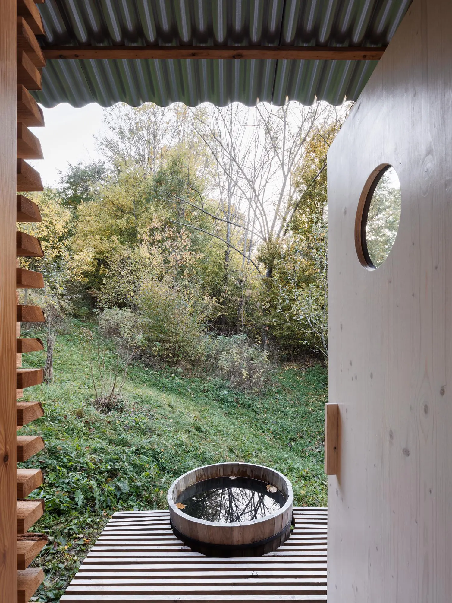 View looking out an open wooden door past a round wooden tub toward a dense green forest valley on a slope.