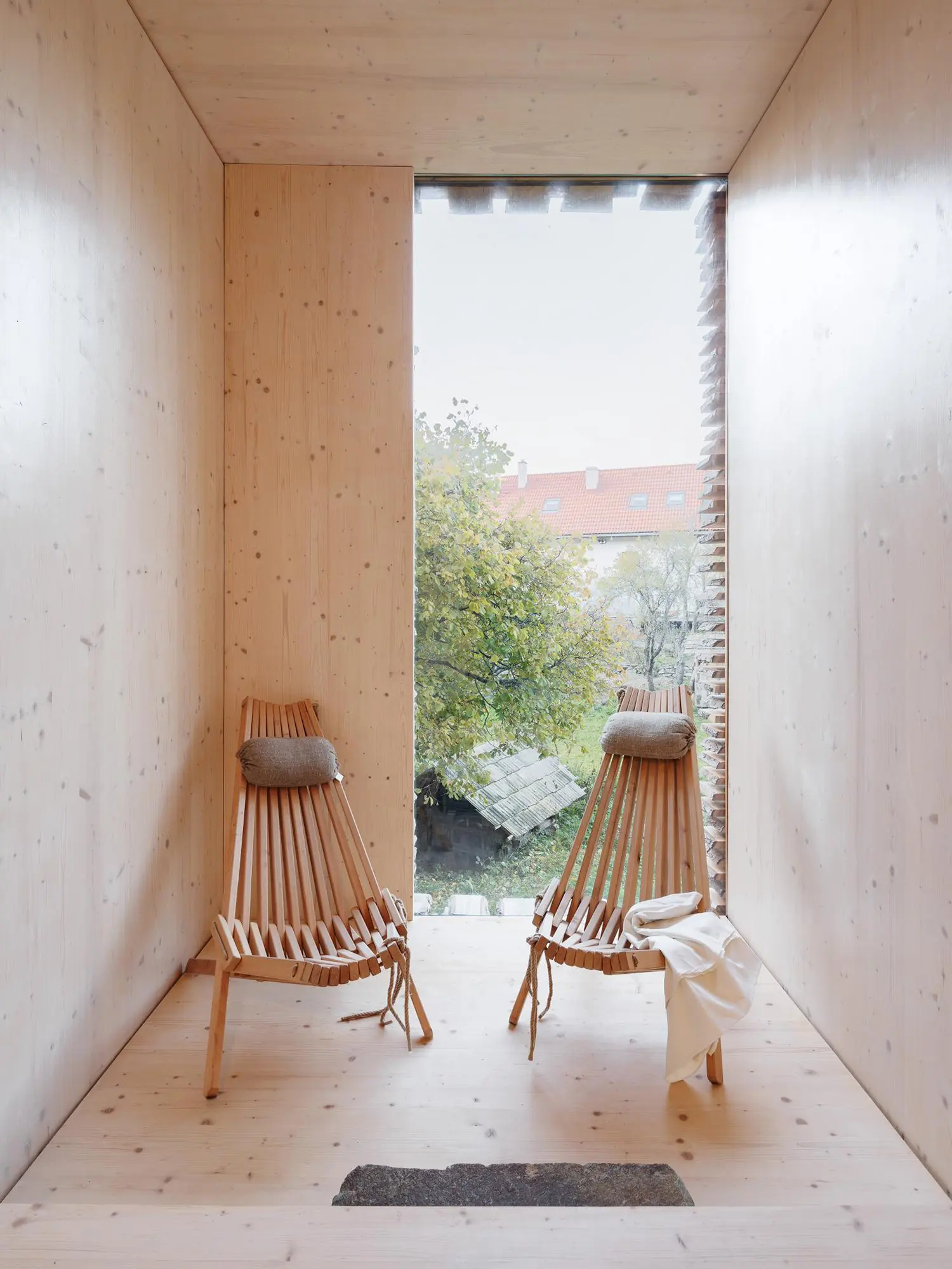 Minimalist spruce-lined relaxation room with two wooden lounge chairs facing a tall window looking out at trees.