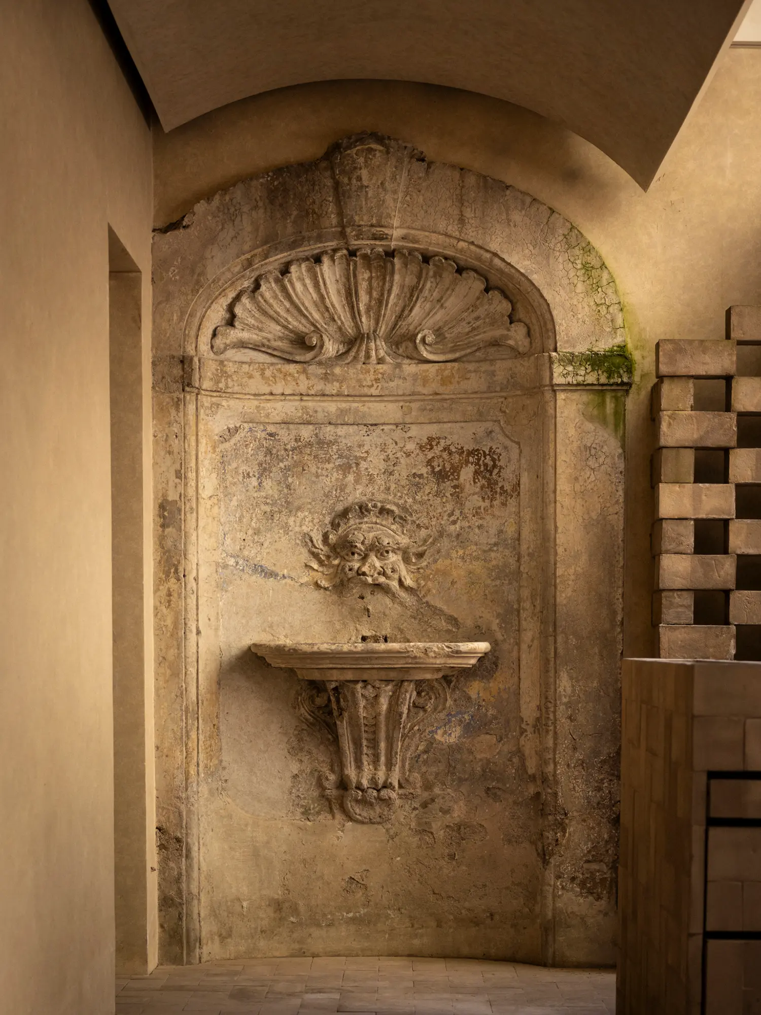 Weathered historic stone wall fountain with a carved face mask inside Nomos Hotel in Rome.