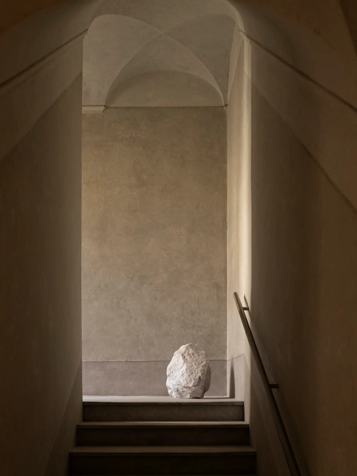 Minimalist stone staircase under a vaulted plaster ceiling featuring a raw block of white marble.