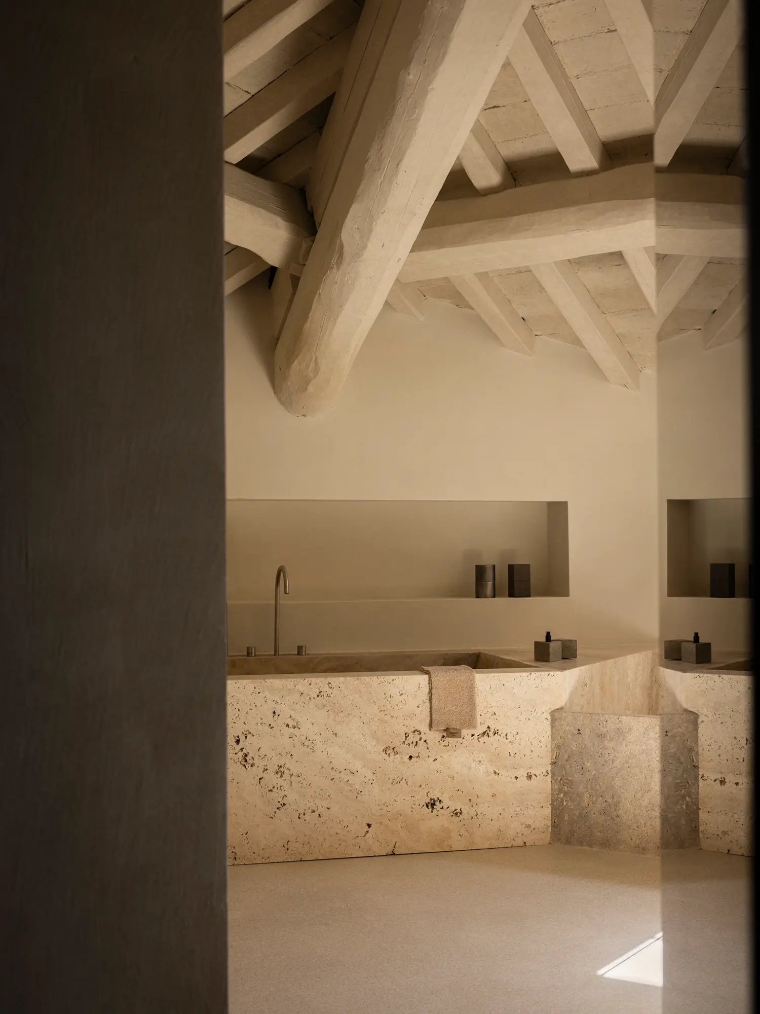 Bathroom at Nomos Hotel featuring a monolithic raw travertine bathtub under exposed timber ceiling beams.