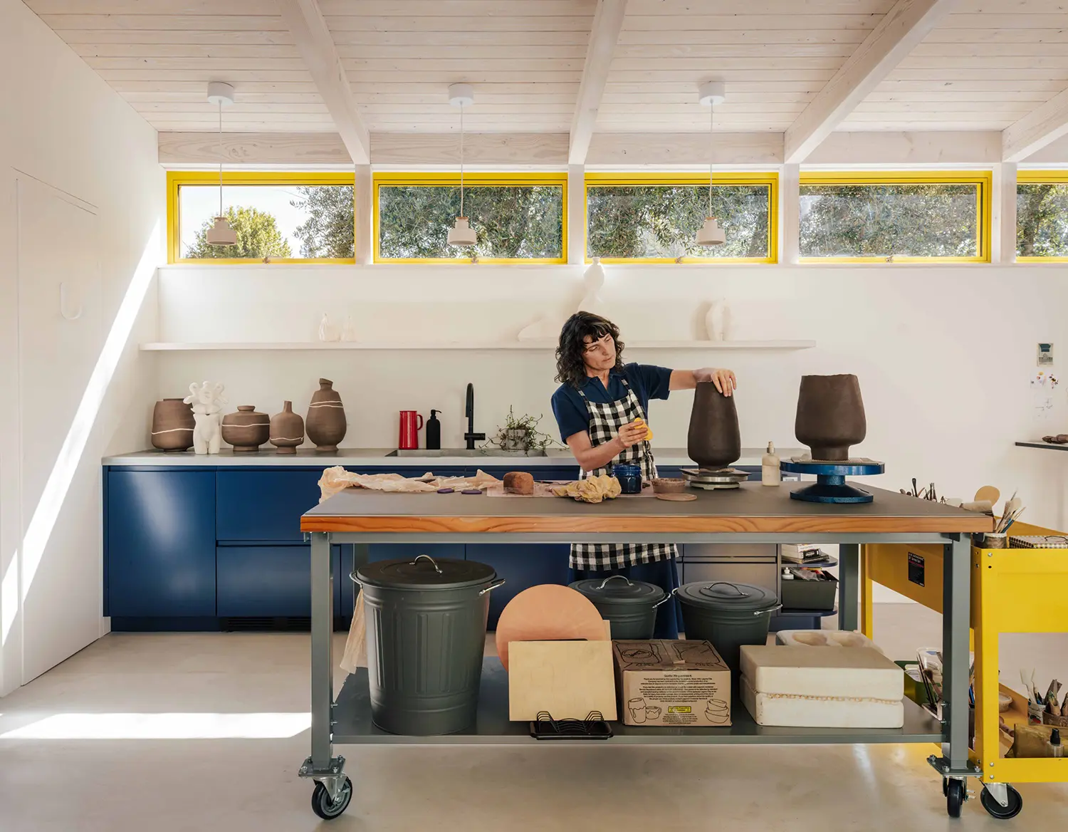 Ceramics artist working at mobile workbench in white garden studio with yellow-framed clerestory windows, Two-Fold Studio Fairfax, ONO