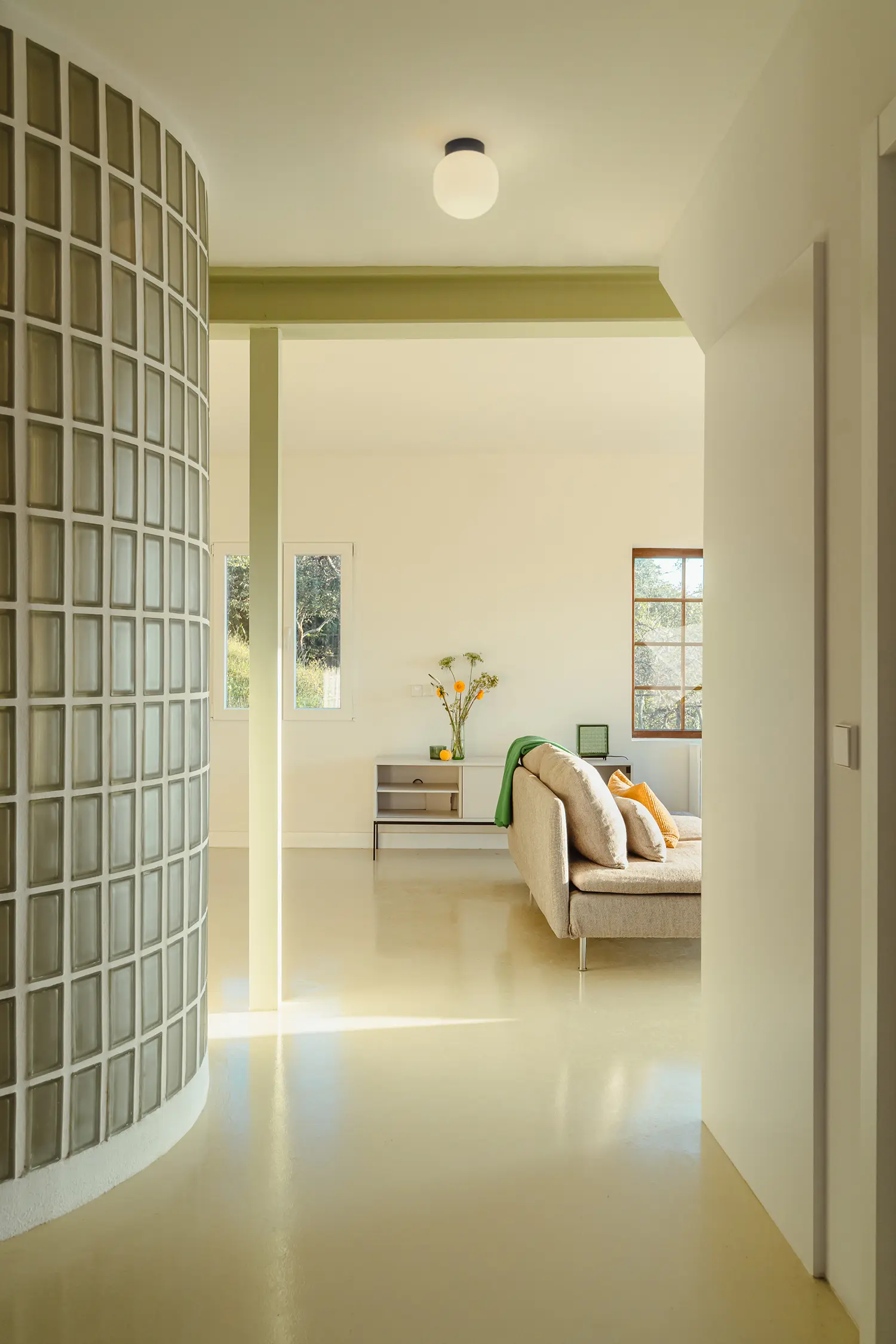 Curved glass-block wall illuminating a minimalist hallway with a beige sofa and pale green structural beams.