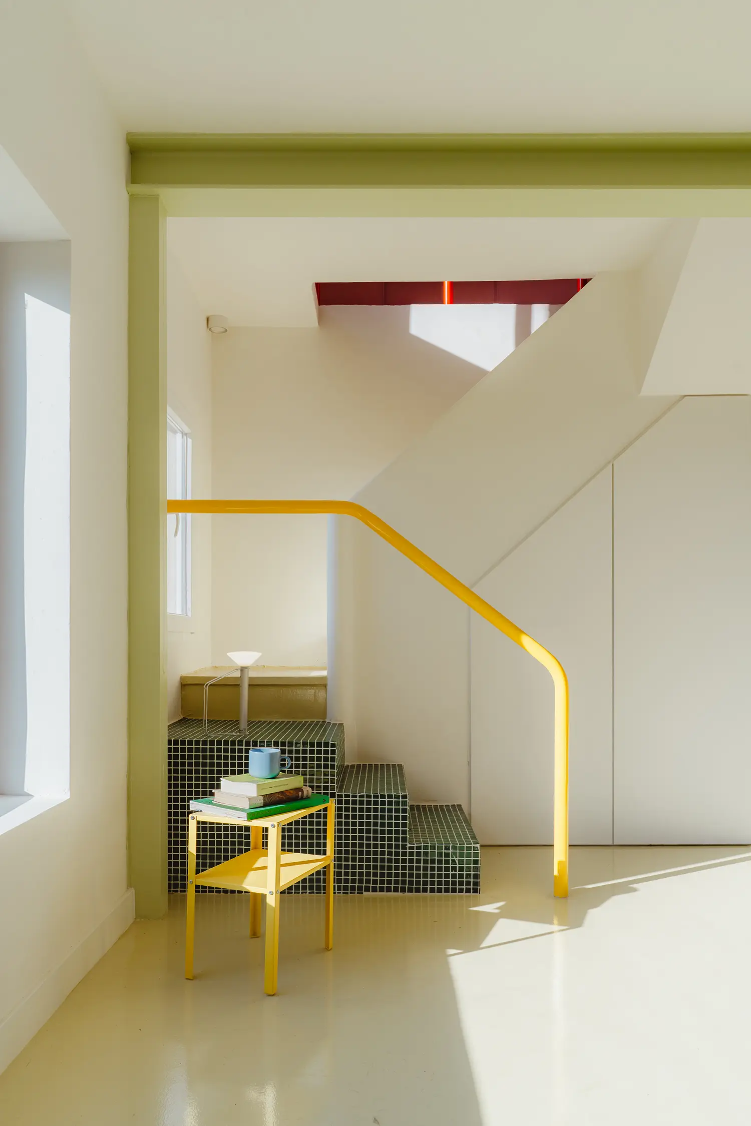 Interior detail of a staircase with green mosaic tiles, a yellow industrial handrail, and a yellow metal side table.
