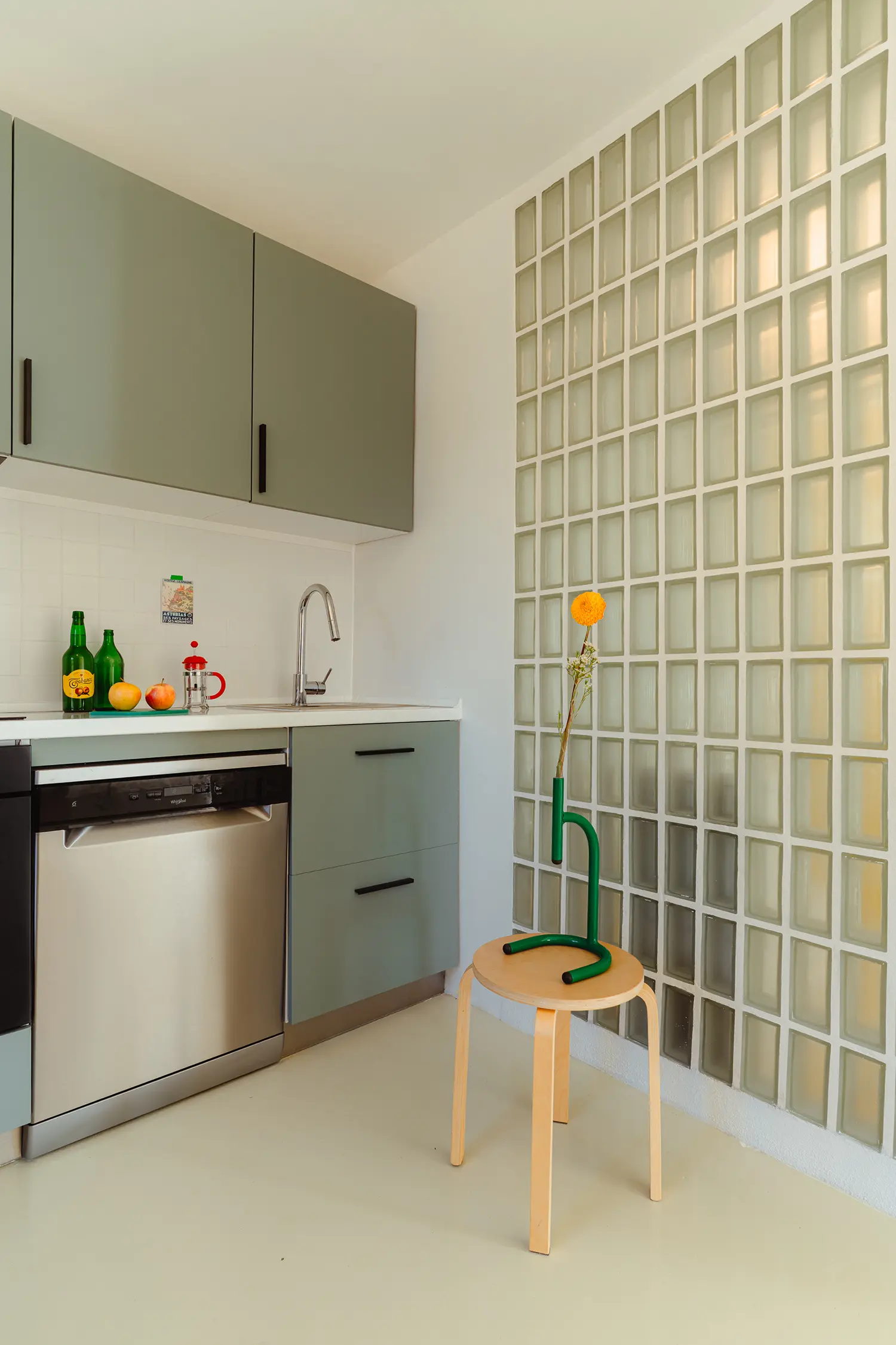 Modern kitchen corner with sage green cabinets next to a glass-block wall and a minimalist wooden stool.
