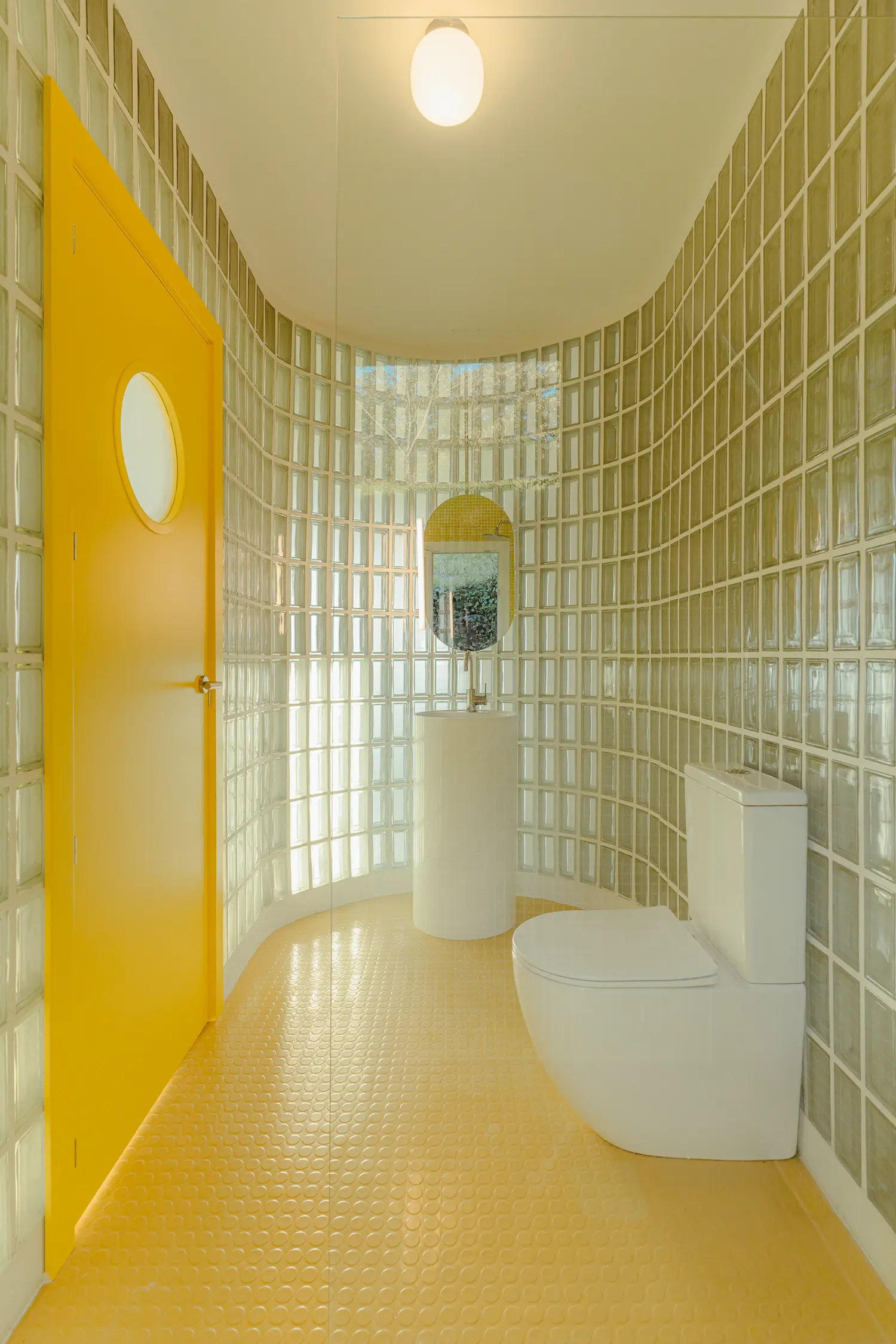 Vibrant yellow bathroom with a circular porthole door, yellow studded rubber flooring, and curved glass-block walls.