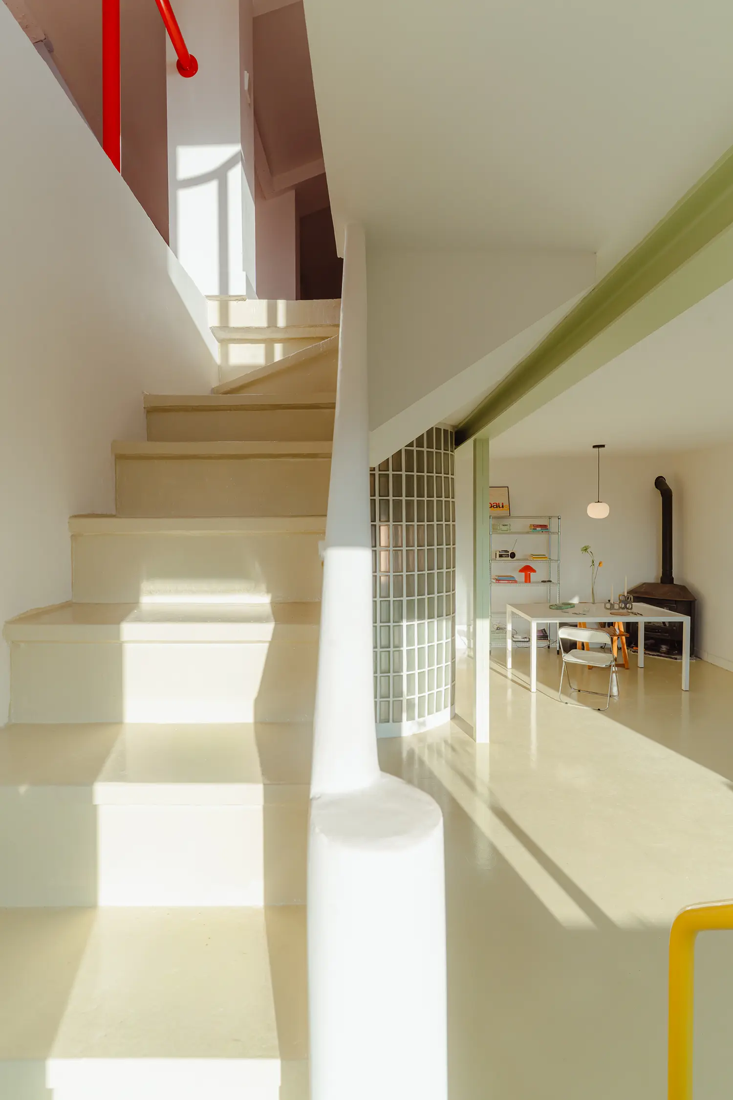 Perspective view from a beige staircase looking toward a dining area with a glass-block volume and a wood-burning stove.