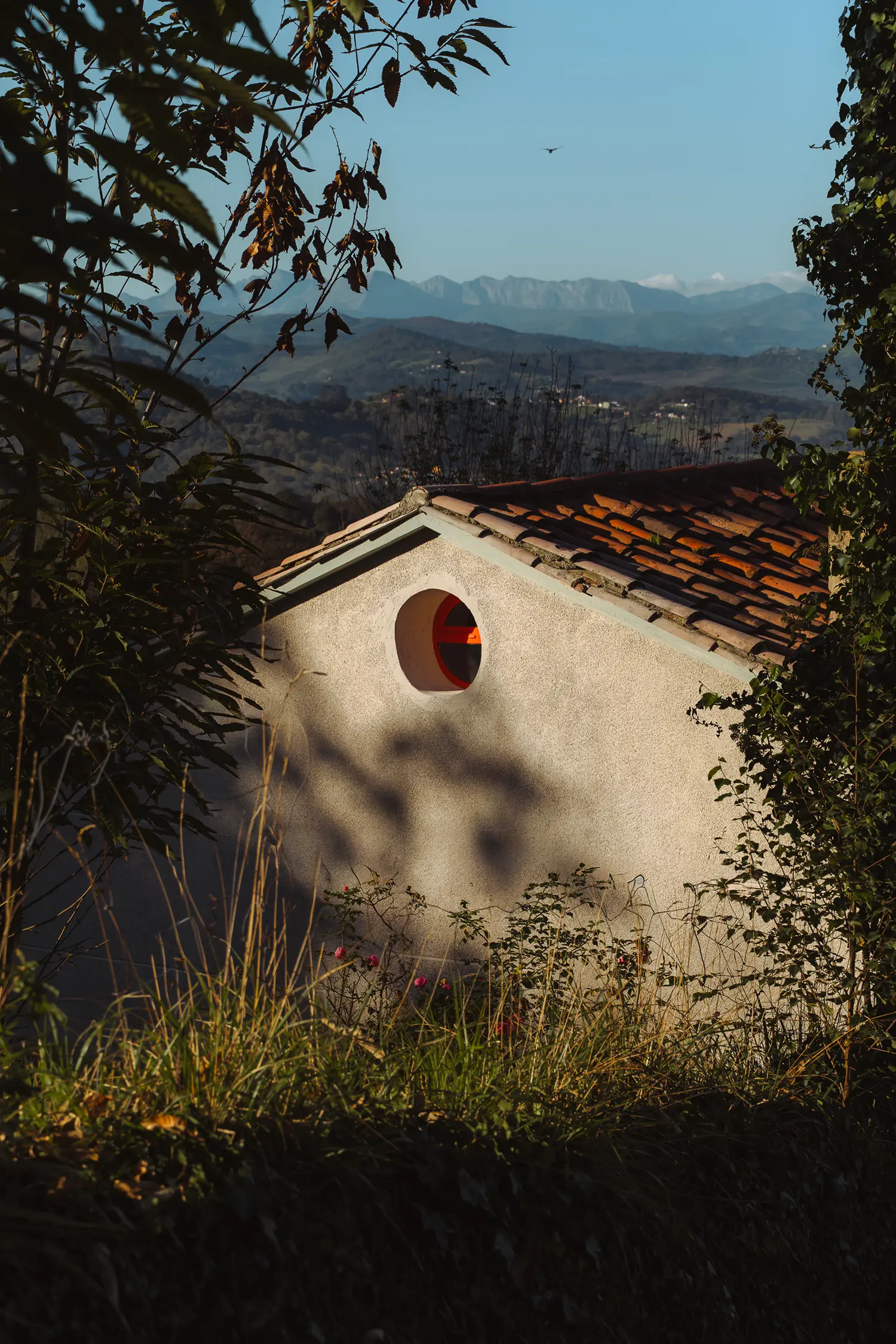 Exterior view of a stone house gable with a circular window showing a red frame against a mountain backdrop.