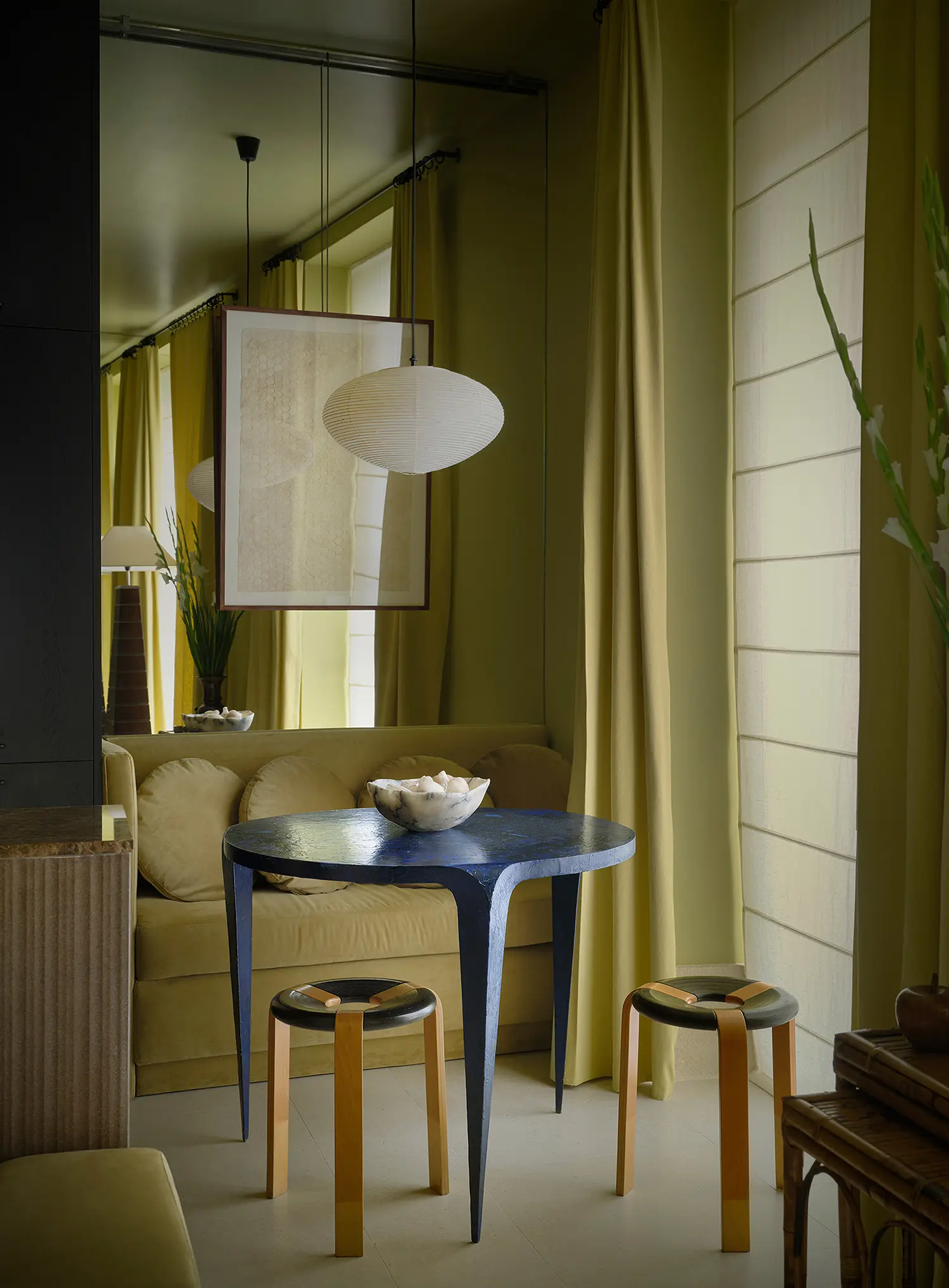 A compact dining corner with a deep blue textured round table, two wooden stools, and a large wall mirror reflecting a classic paper pendant lamp.