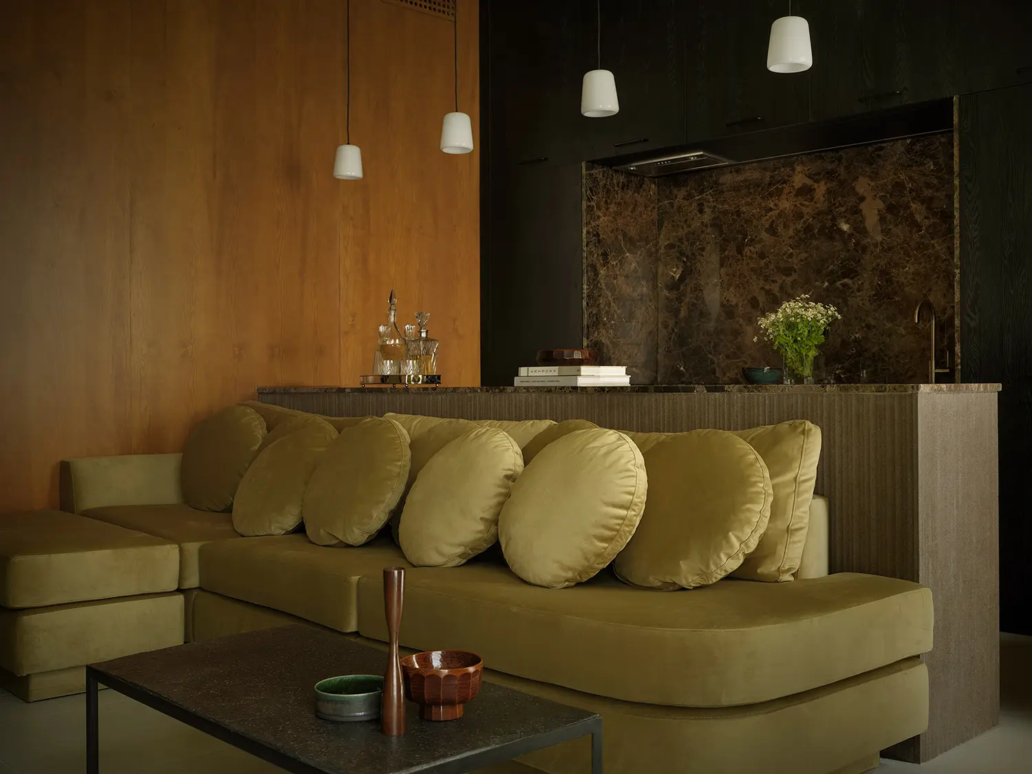 Low olive velvet sofa with round bolster cushions backed by a fluted stone kitchen counter under minimalist white pendant lights.