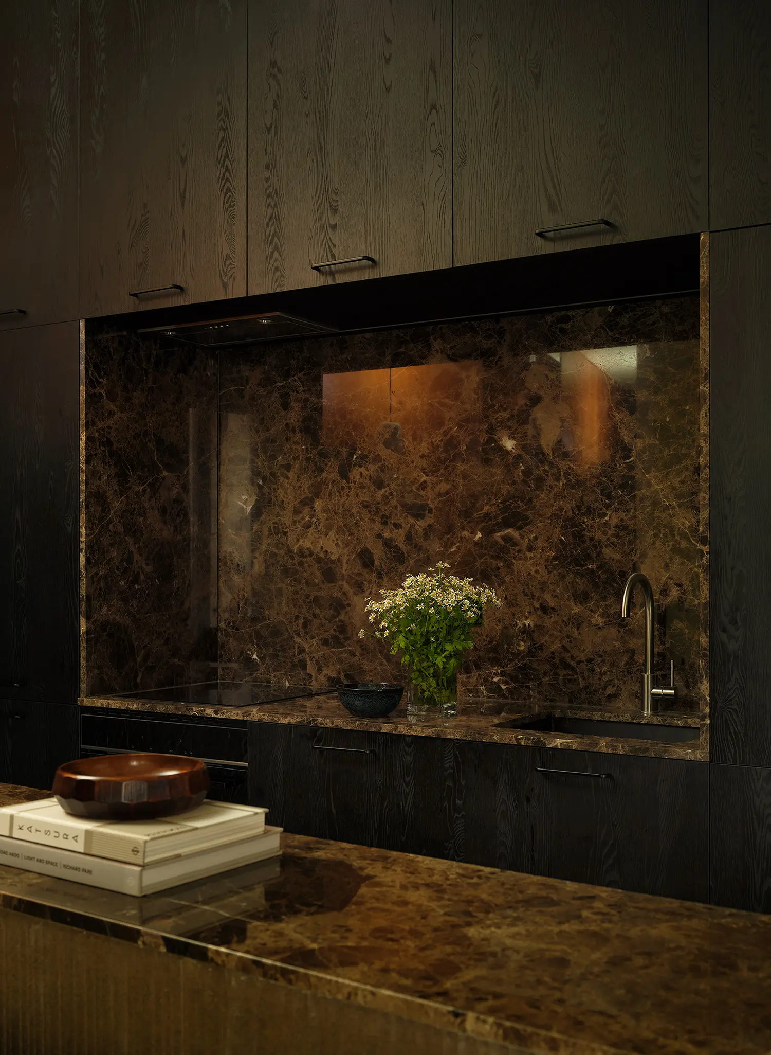 Detailed close-up of a monolithic dark wood kitchen niche with an integrated cooktop, sink, and polished Emperador brown marble surfaces.