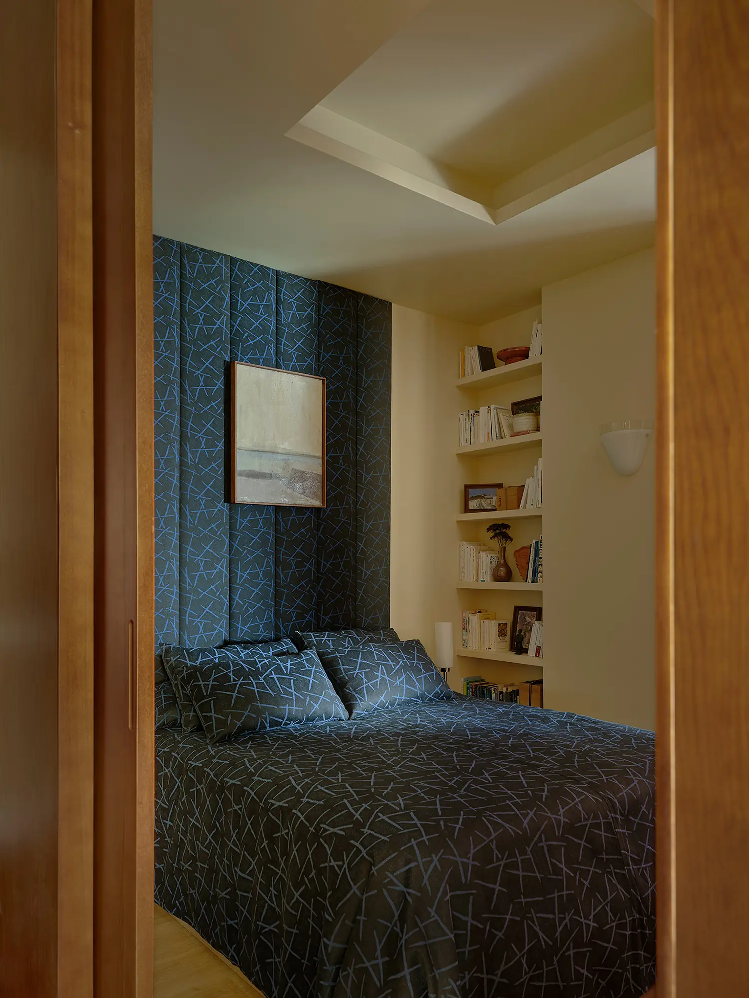 Intimate bedroom interior viewed through an oak doorway, showcasing a dark blue patterned headboard panel and built-in white bookshelves.