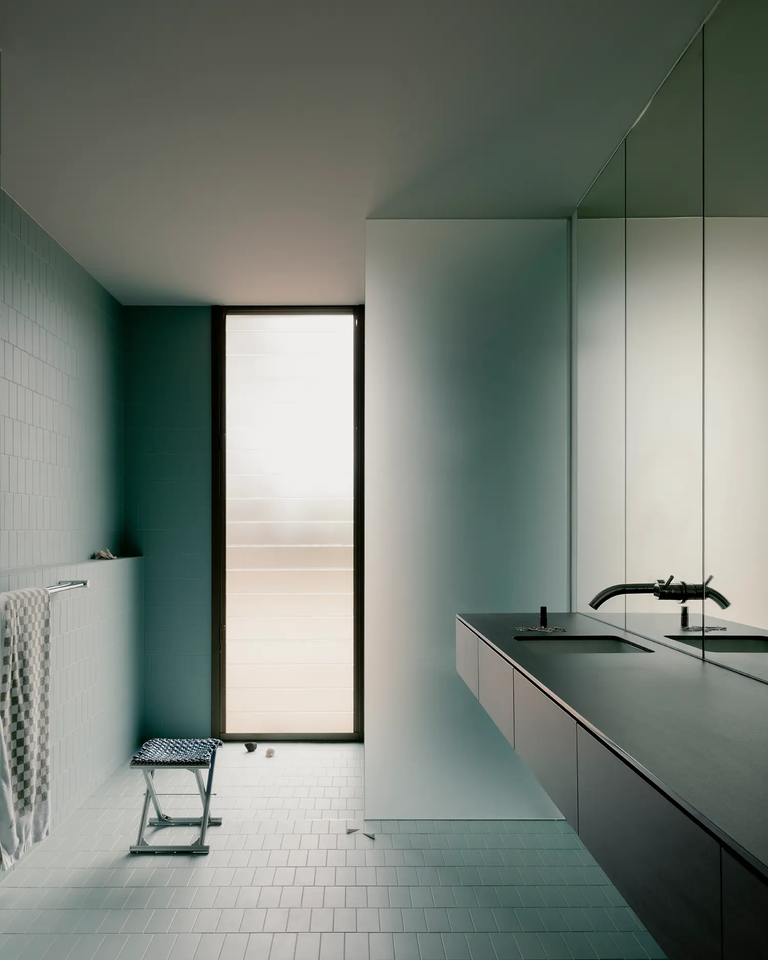Minimalist bathroom design with pale green tiles, a long black vanity, and a large frosted window.