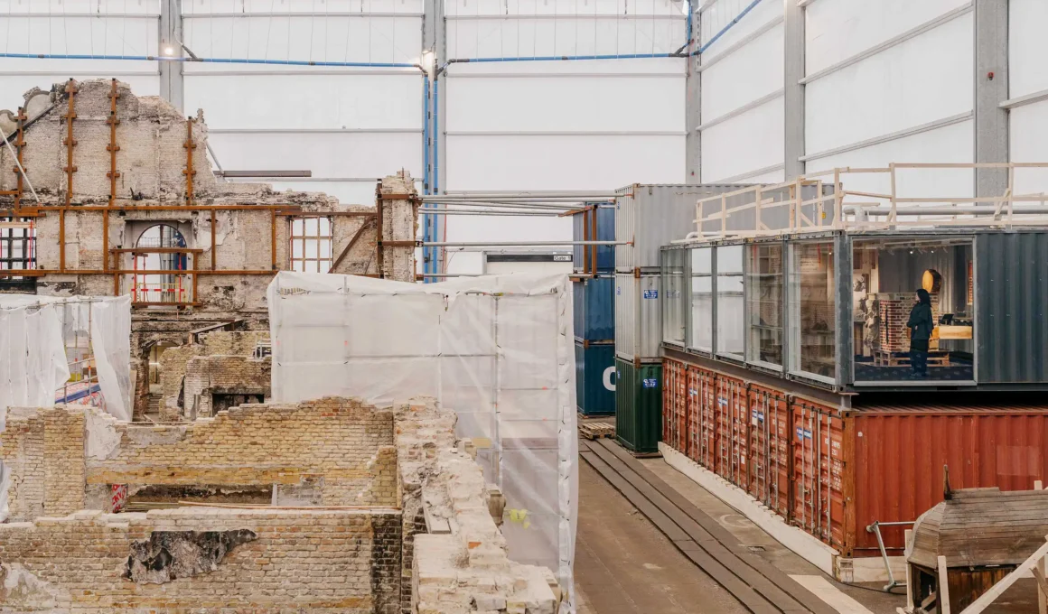 Stacked industrial shipping containers positioned next to the exposed brick ruins of the Børsen building under a white protective canopy.