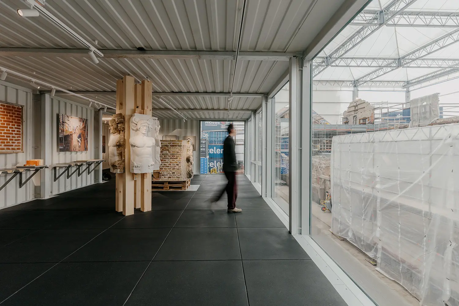 Interior of a grey shipping container gallery featuring large glass windows overlooking a construction site and classical white bust sculptures mounted on timber columns.