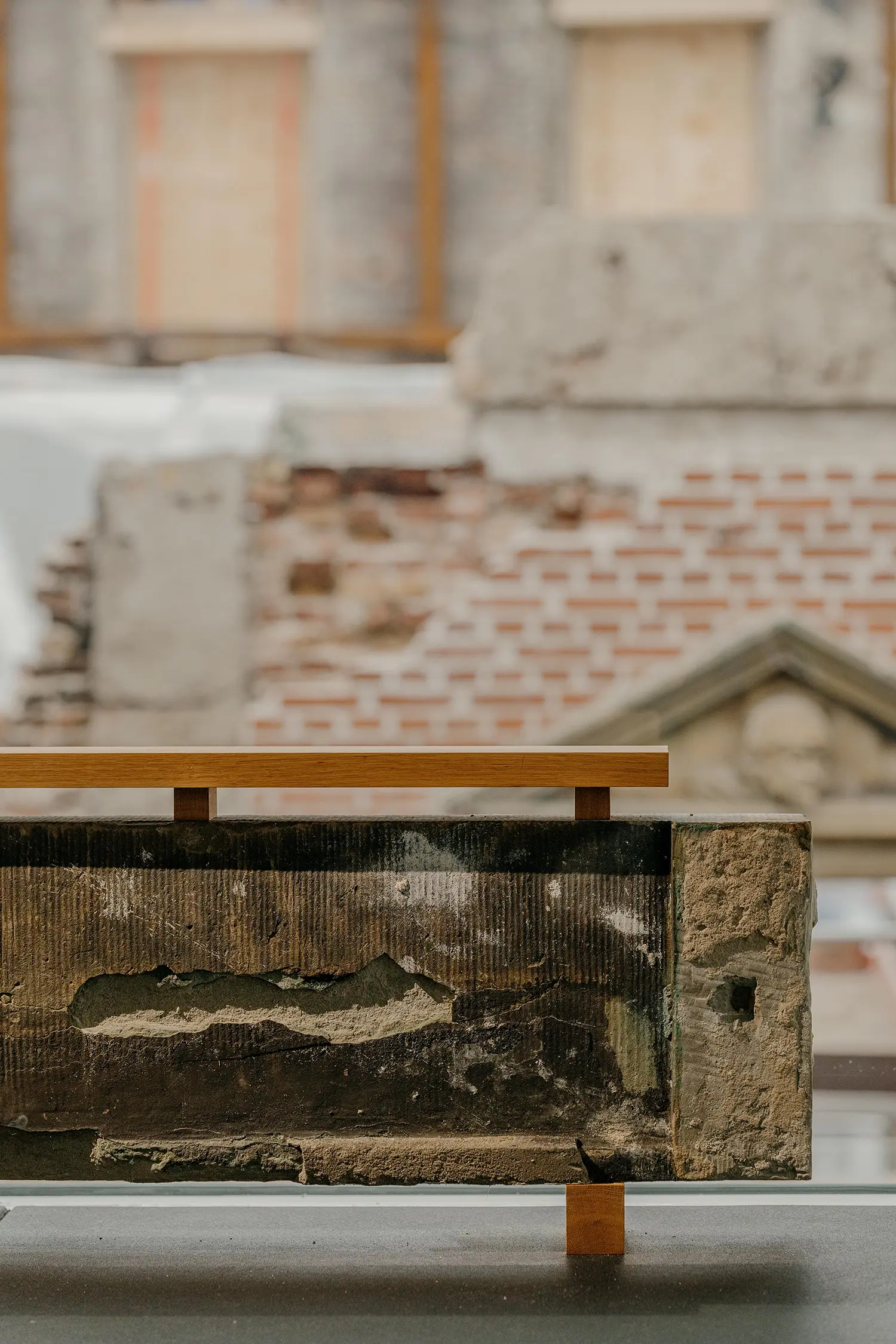 Close-up of a weathered stone architectural fragment mounted on a wooden stand with the blurred brickwork of Børsen in the background.