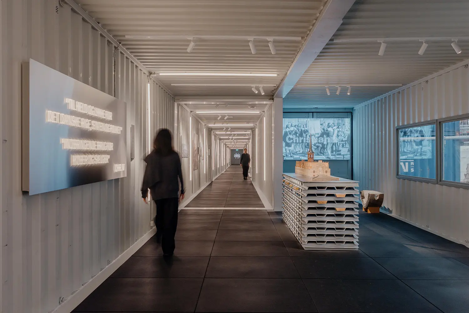 Long perspective of a brightly lit corridor made of interconnected white shipping containers with a scale model of Børsen in the center.