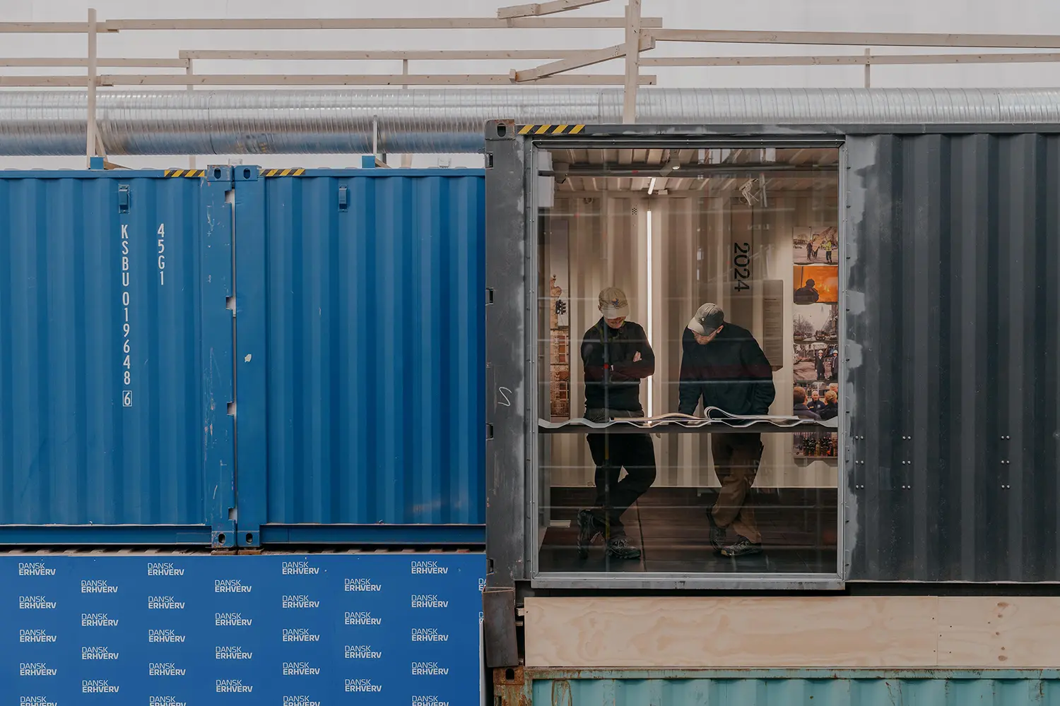 Two people inside a grey shipping container looking at architectural plans near a blue container marked with Dansk Erhverv logos.