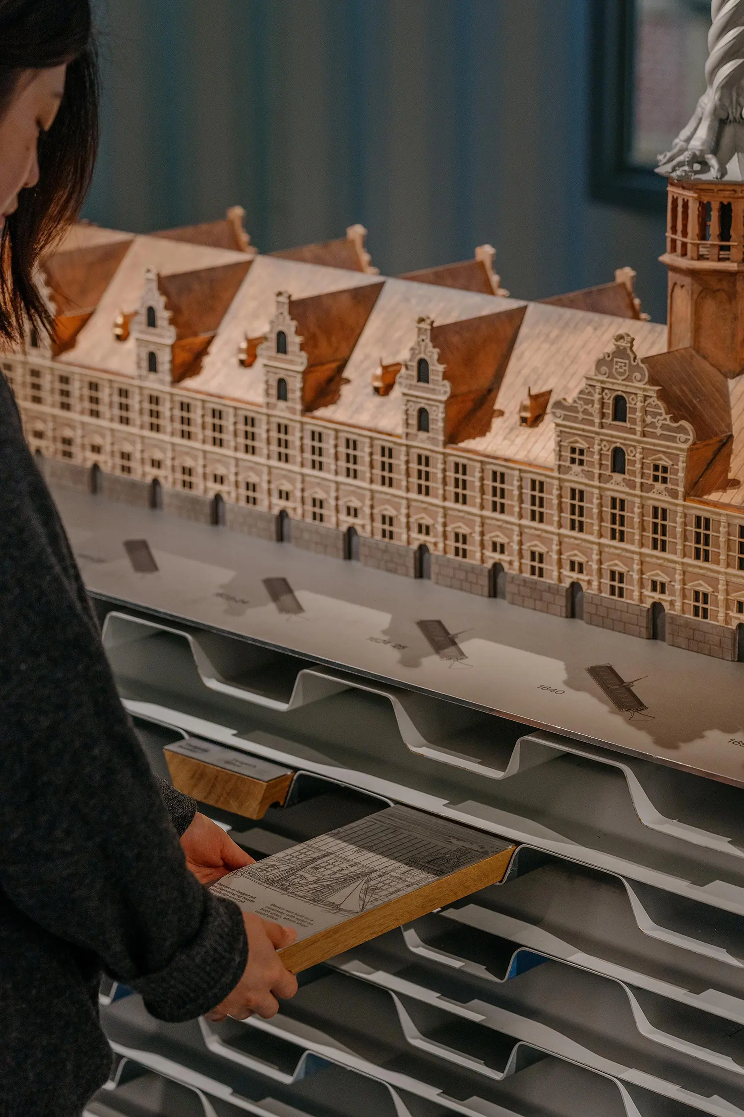 A person pulling a wooden drawer containing an architectural drawing from a base made of stacked corrugated metal sheets.