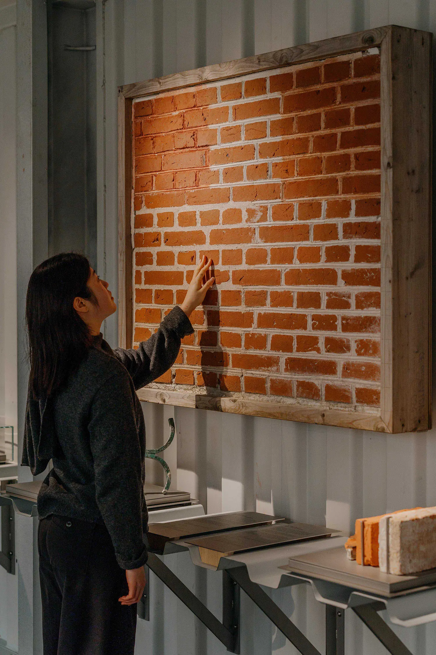 A woman touching a framed section of red brick wall mounted inside a corrugated metal gallery space.