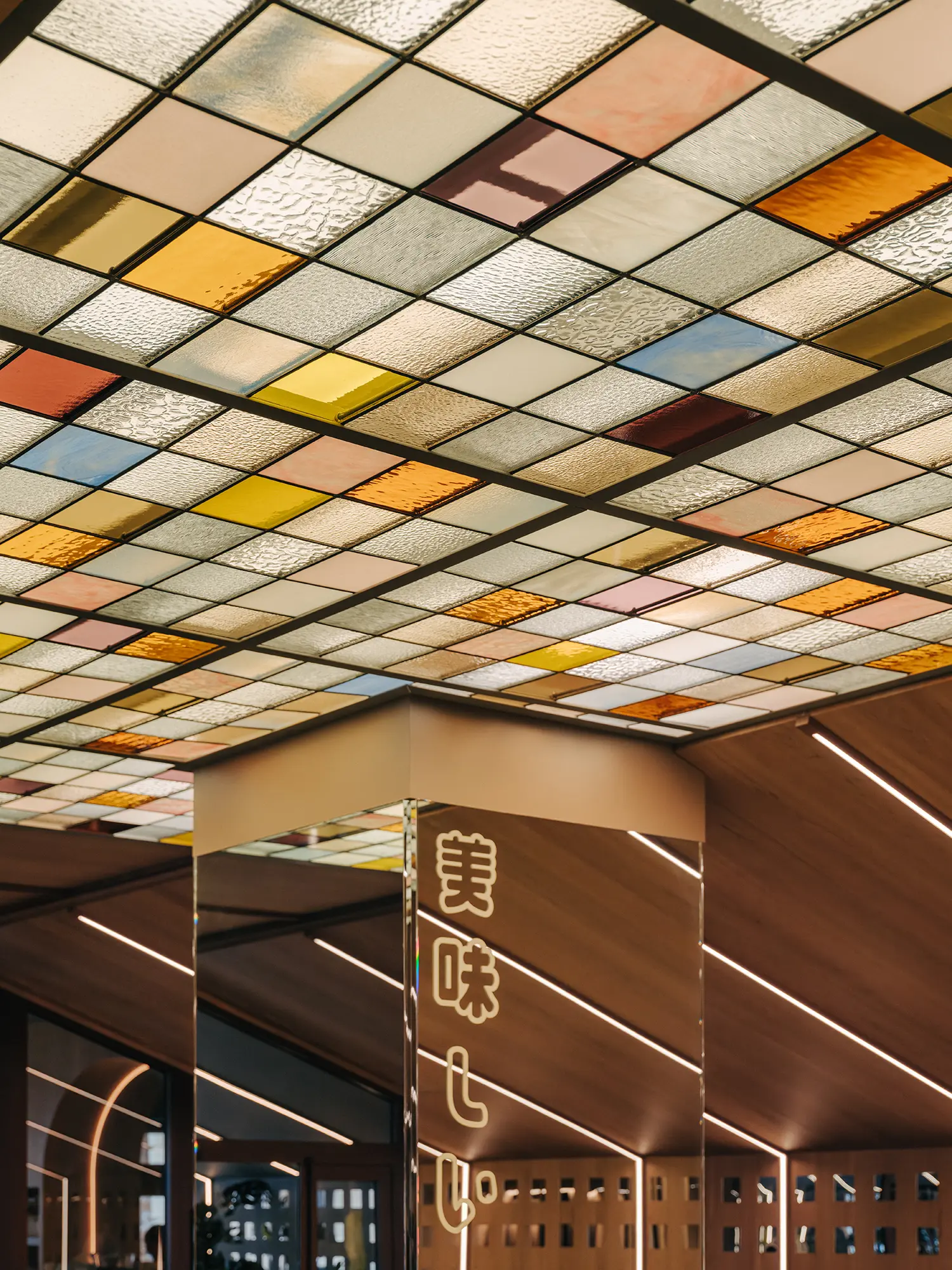 Architectural detail of a gridded, pastel-colored stained-glass ceiling and a mirrored pillar with illuminated Japanese neon writing in Matsuri Marseille.