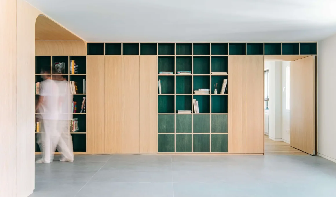 Custom oak cabinetry wall with deep green open shelving grids and grey large-format tile flooring in a Madrid apartment.