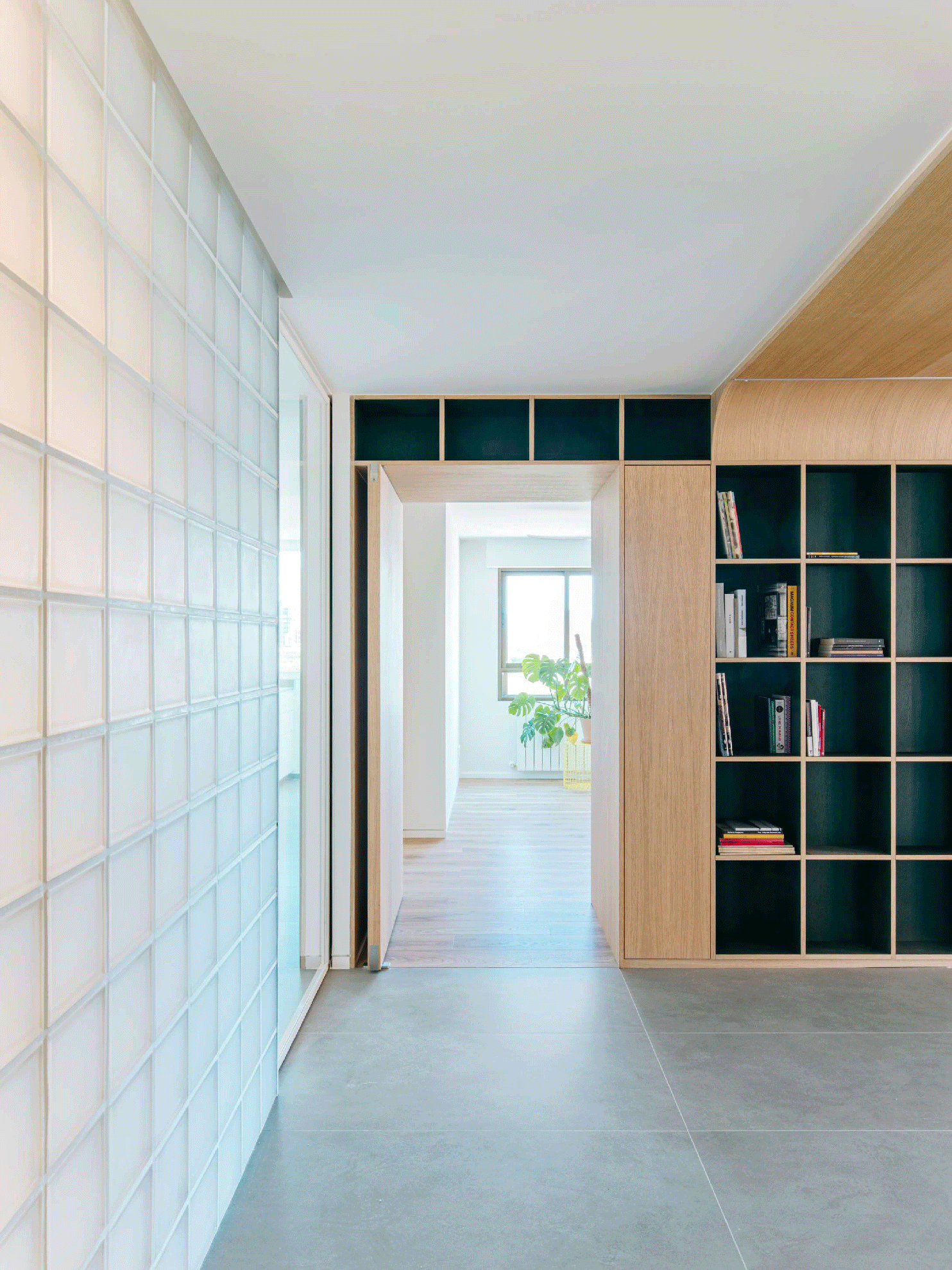 Translucent glass-block partition wall framing a view past a custom oak shelving unit toward a sunlit room.