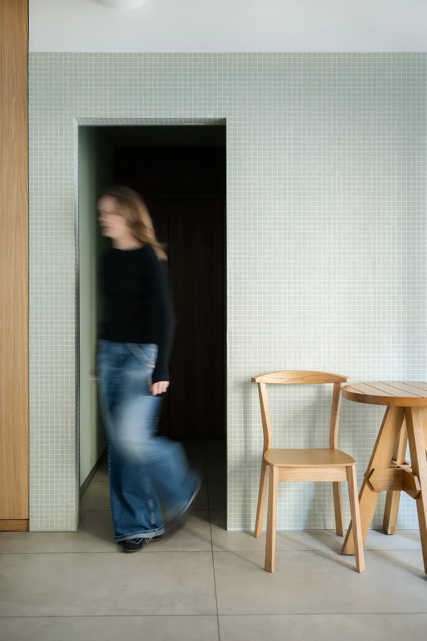 Minimalist doorway framed by light green square wall tiles next to a wooden chair and table.
