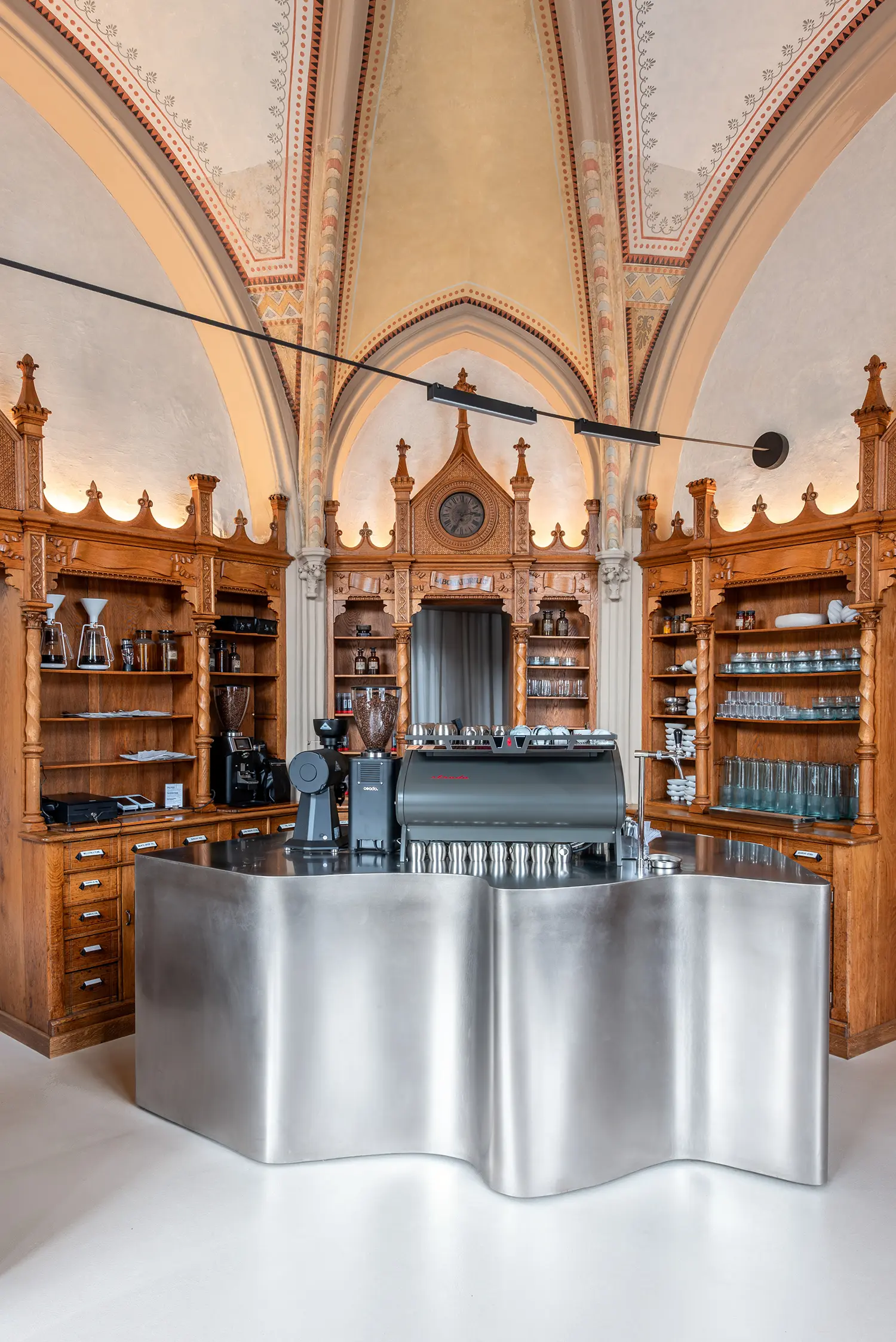 Close-up of a curved stainless steel coffee bar with a professional espresso machine and coffee grinders under a vaulted ceiling.