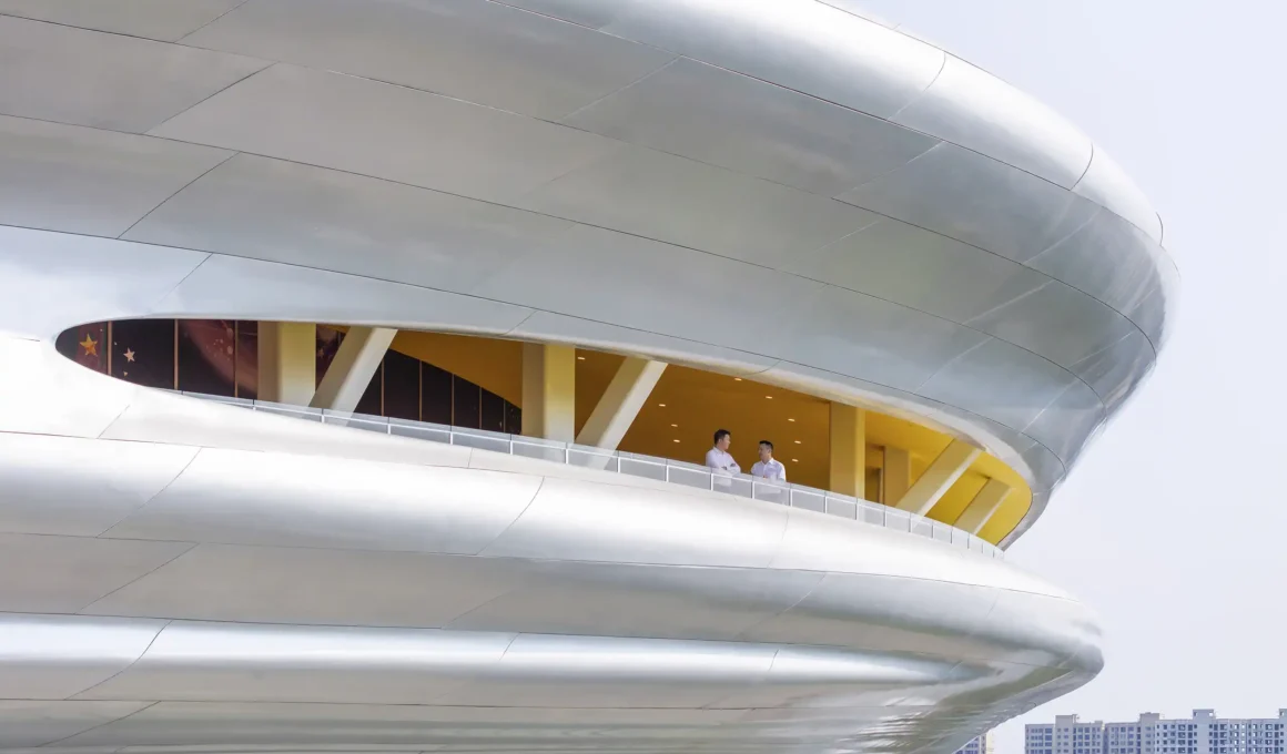 Close-up of the shimmering silver facade of the Hainan Science Museum with two people on an observation balcony.