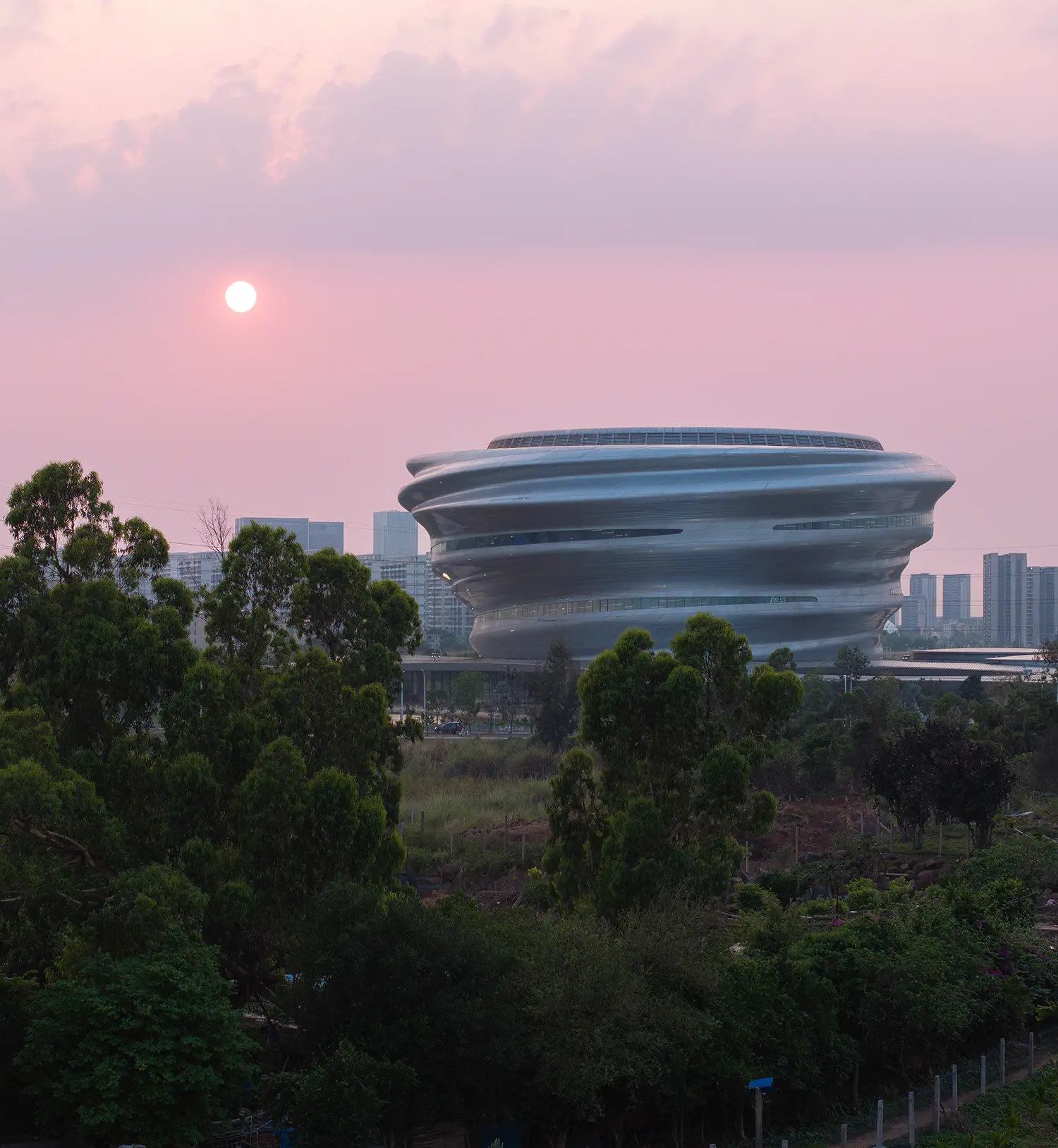 The Hainan Science Museum at dusk with a pink sky and a setting sun behind tropical trees.