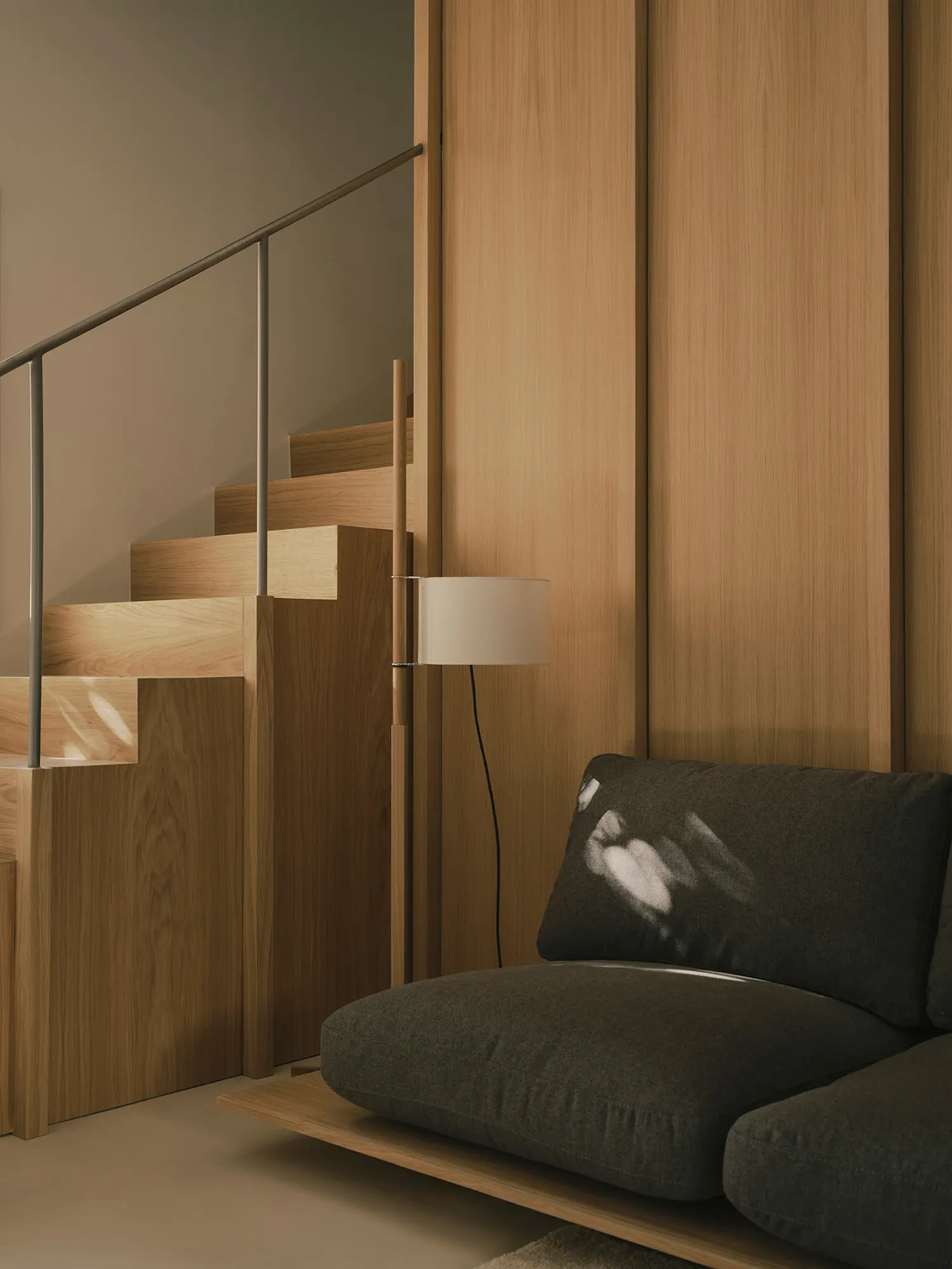 Close-up of an oak staircase next to a dark gray sofa and a minimalist floor lamp.