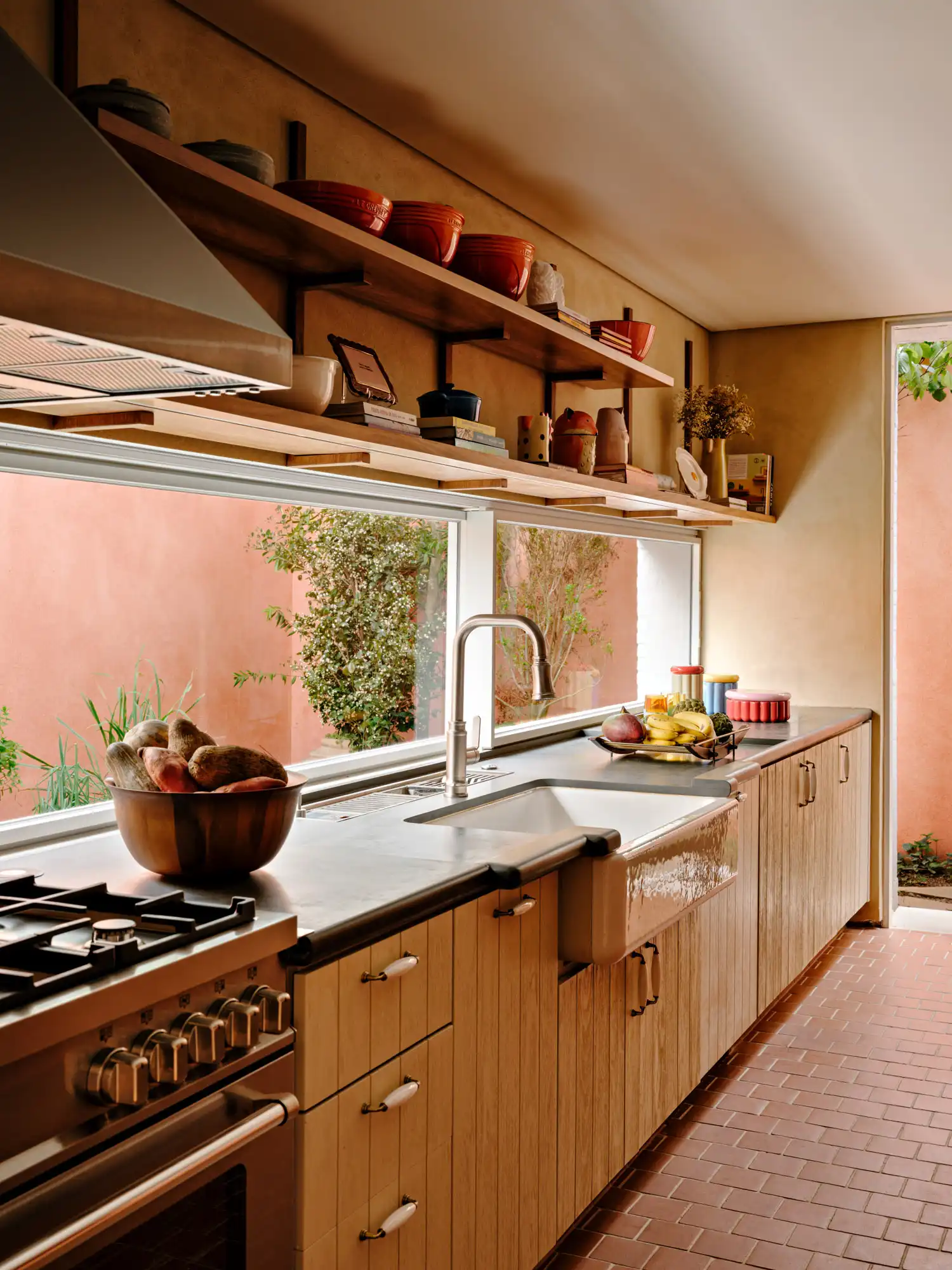 Kitchen with wooden cabinets, reddish countertops, apron sink, tile floor, and a long window facing a terracotta wall.
