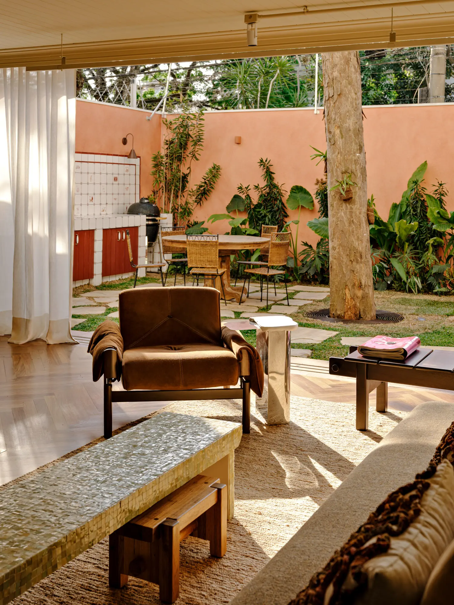 Suede armchair and tiled coffee table in an open lounge looking onto a courtyard with stone paving and a mature tree.