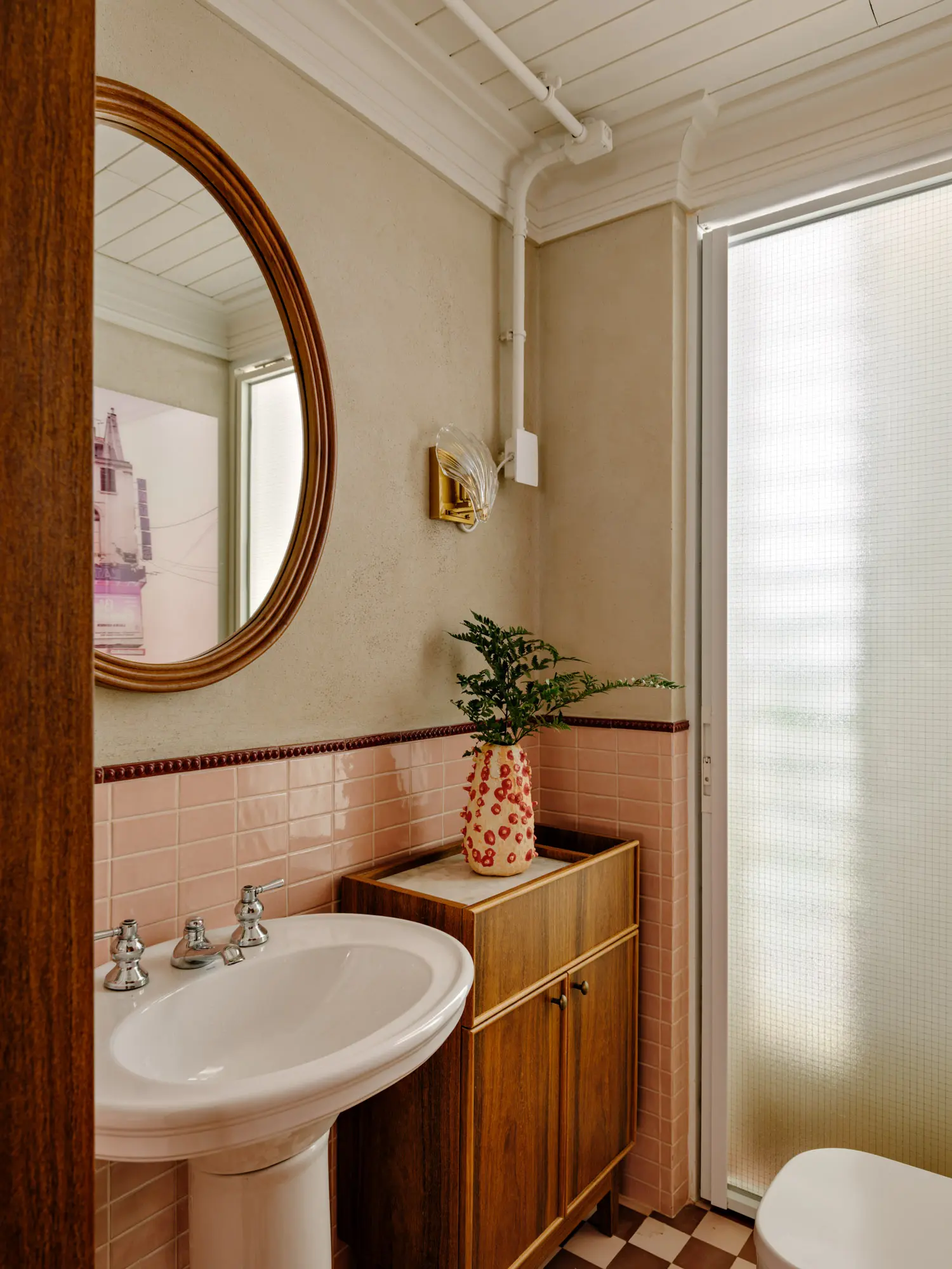 Powder room with a white pedestal sink, wooden mirror, pink wall tiles, and a brown and white checkerboard floor.