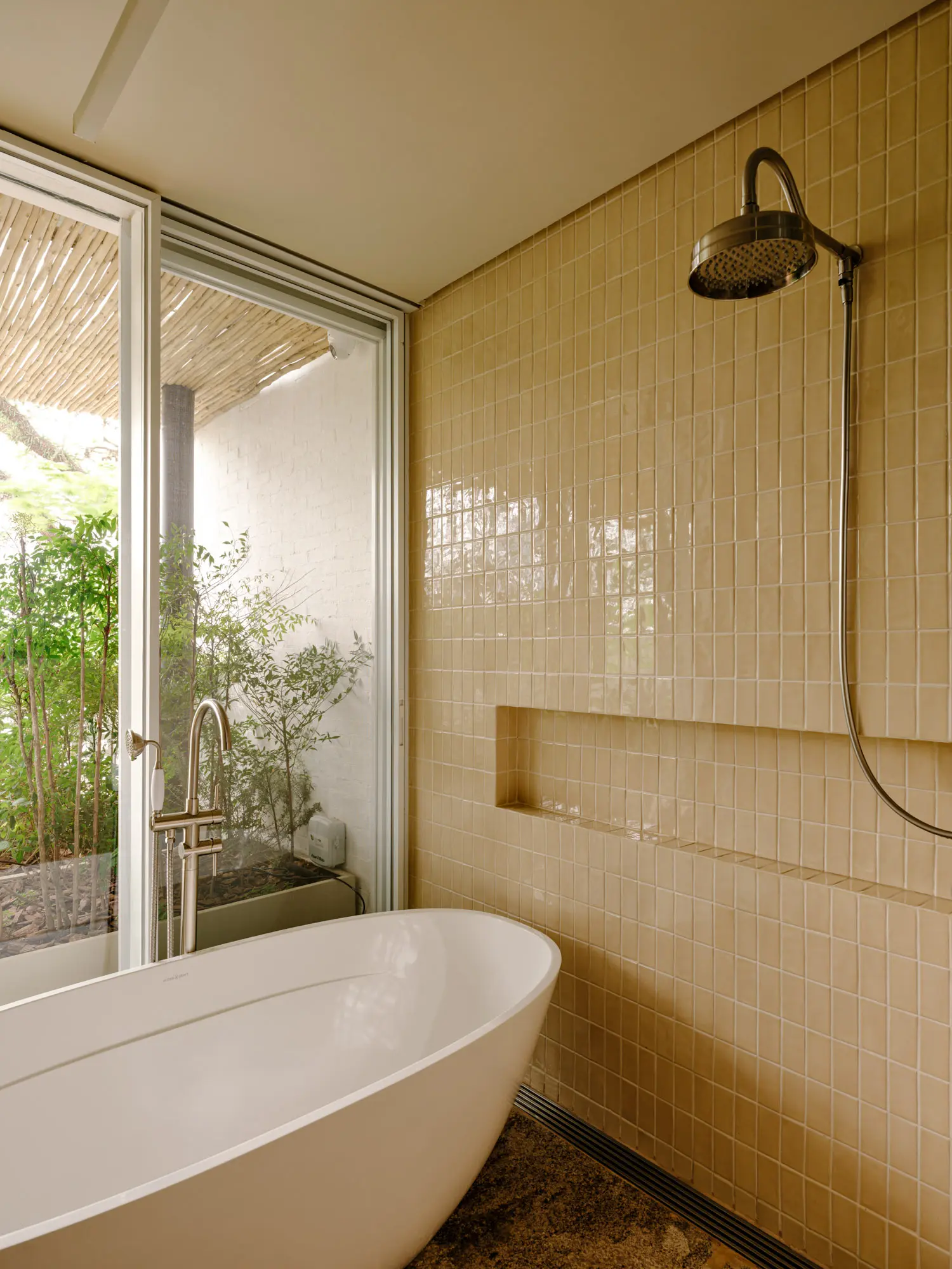 Freestanding white bathtub next to a Canary-yellow tiled wall and a large sliding glass door looking onto foliage.