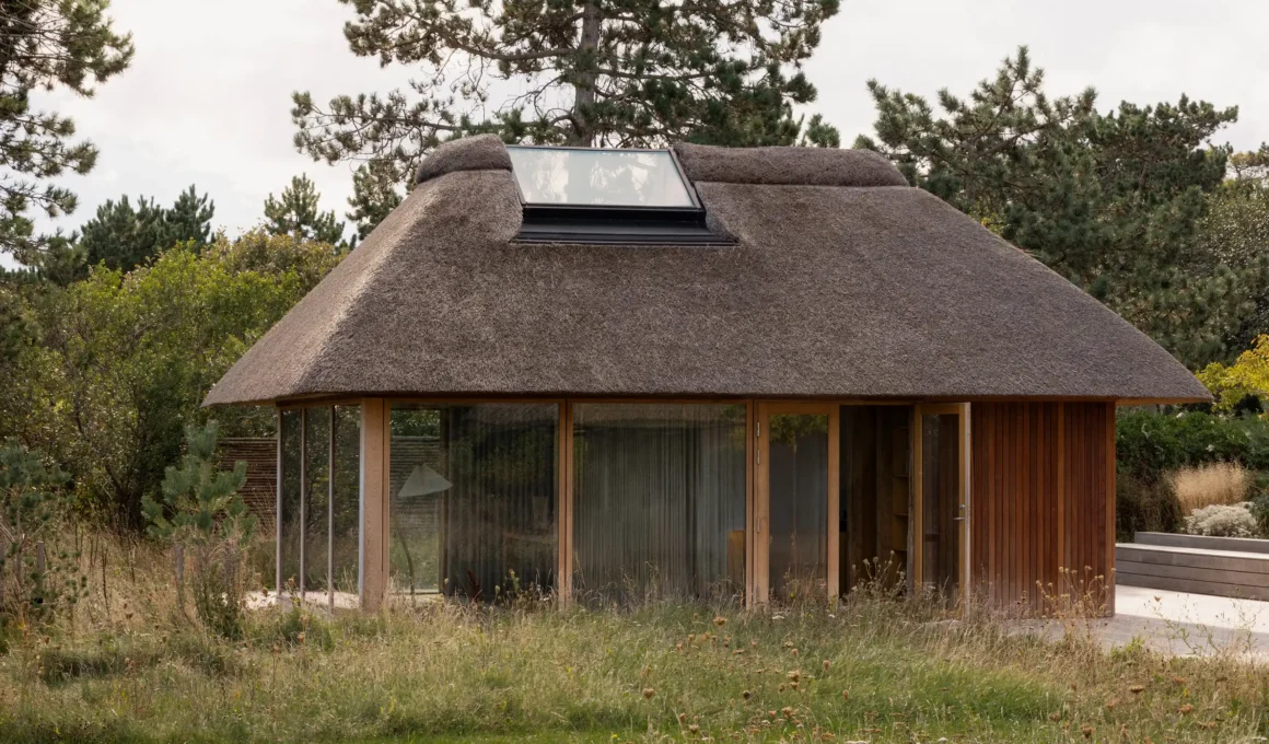 Exterior view of a minimalist wooden pavilion with a thick thatched roof and large glass windows under a clear sky.