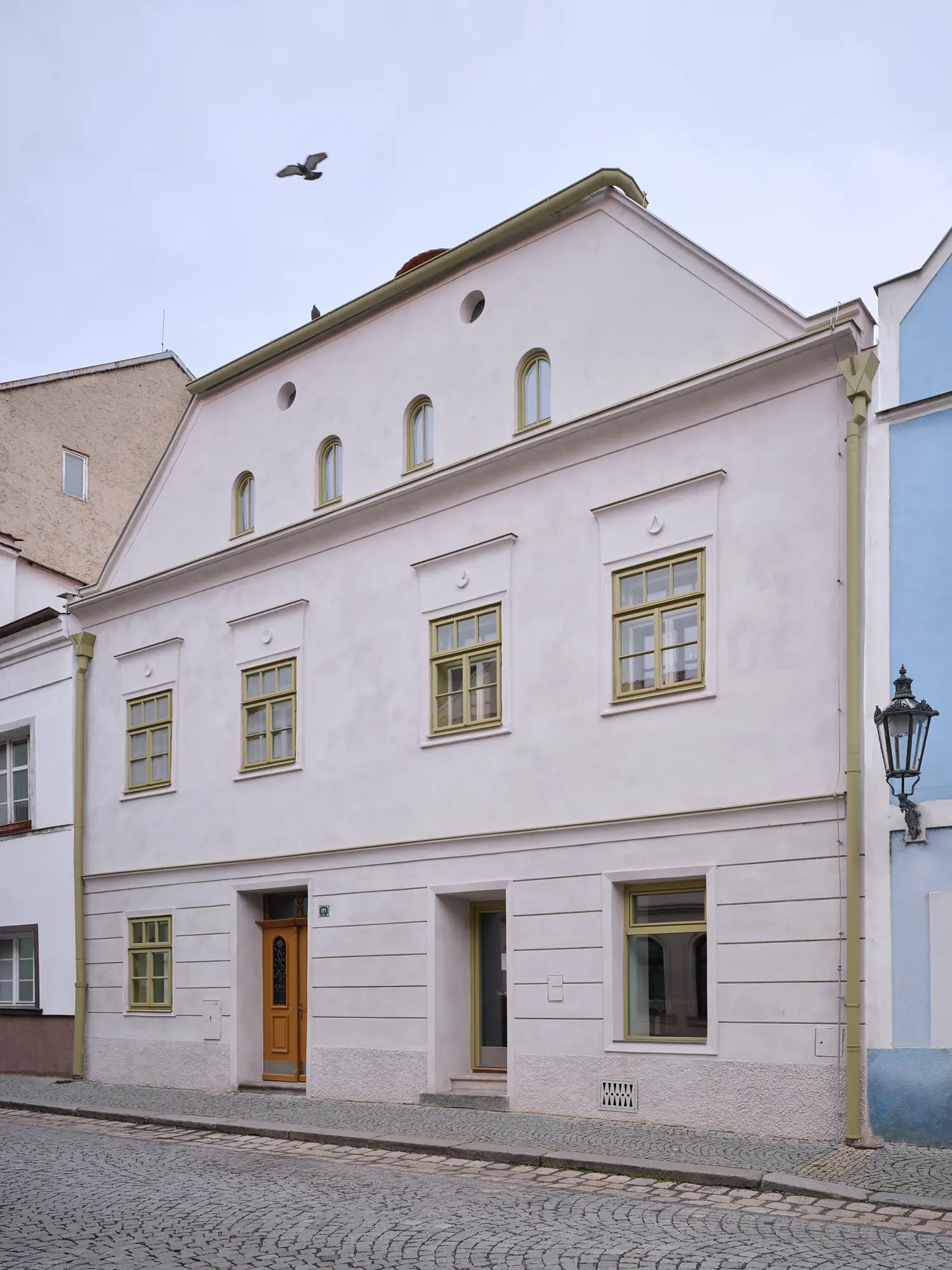 Oblouková 171 ORA — restored street facade with reinterpreted stucco cornice, arched attic windows, yellow-green timber frames, cobblestone street