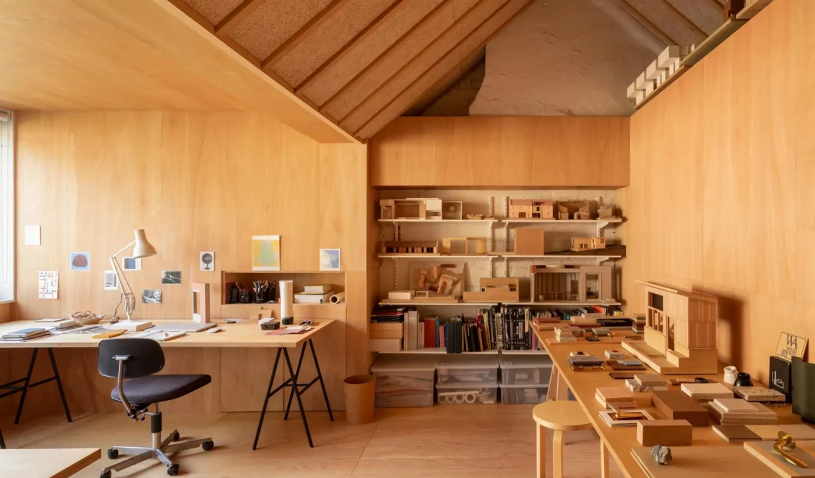 Plywood-lined attic studio with pitched roof, exposed rafters, architectural models, wooden desks, and an office chair.